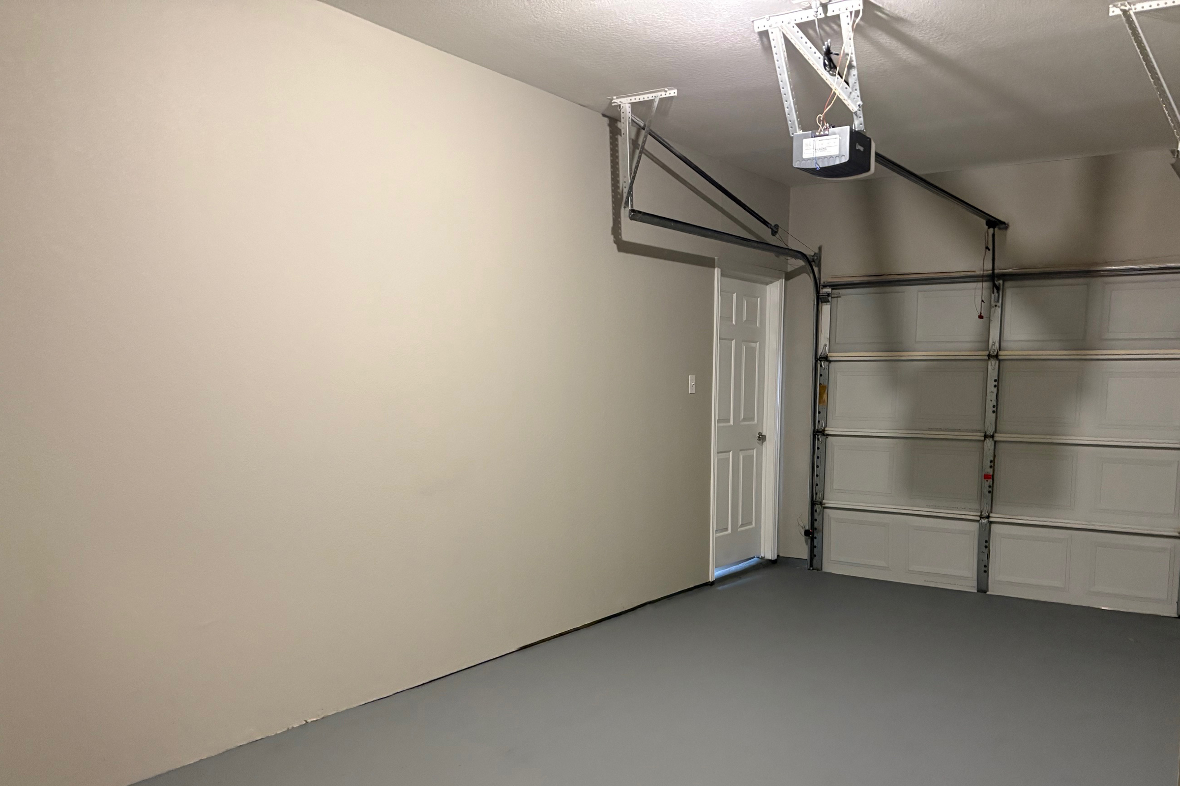 A clean and empty garage space featuring a light gray wall, a closed door on the left, and a white garage door made of sectional panels on the right. The floor is painted in a light gray color, and overhead, there are metal tracks for garage door operation. The overall atmosphere is tidy and organized.