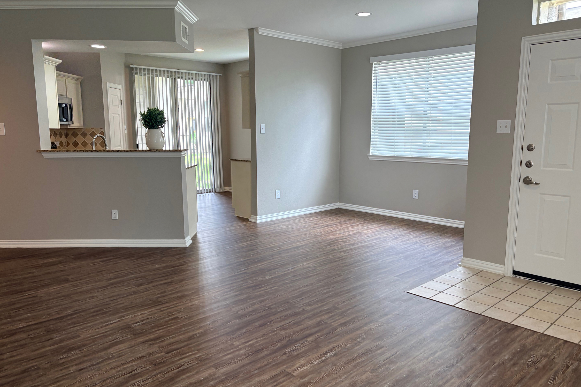 An interior view of a spacious, empty living room with wooden laminate flooring and neutral-colored walls. A small entryway with a tiled floor is visible to the right, and a kitchen area is seen in the background. Natural light enters through the window, illuminating the space.