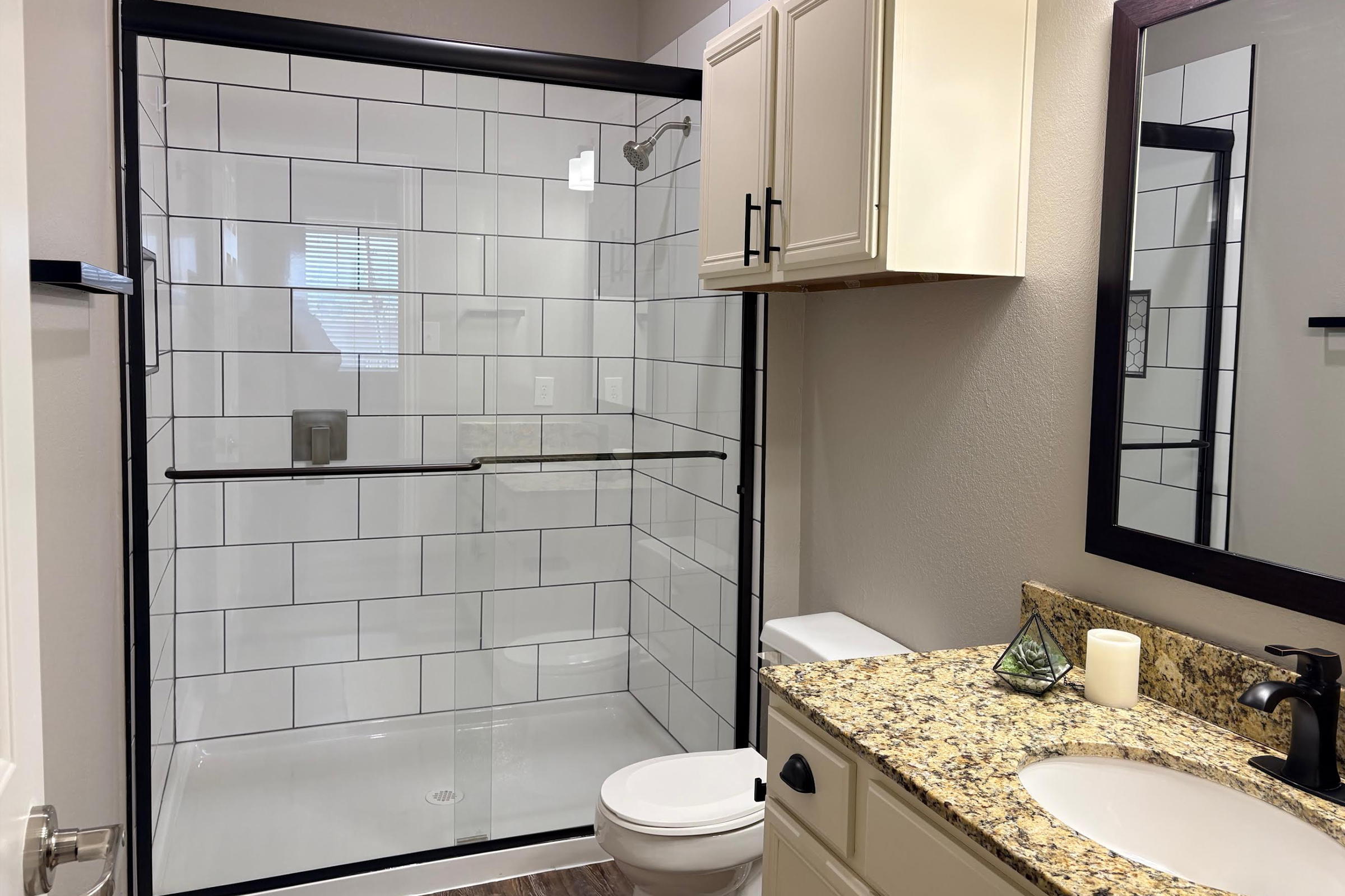A modern bathroom featuring a walk-in glass shower with tile accents, a granite countertop with a sink, a mirror, and a toilet. The walls are painted in neutral tones, and there is a small decorative item on the counter. Natural light comes in through a window in the shower area.