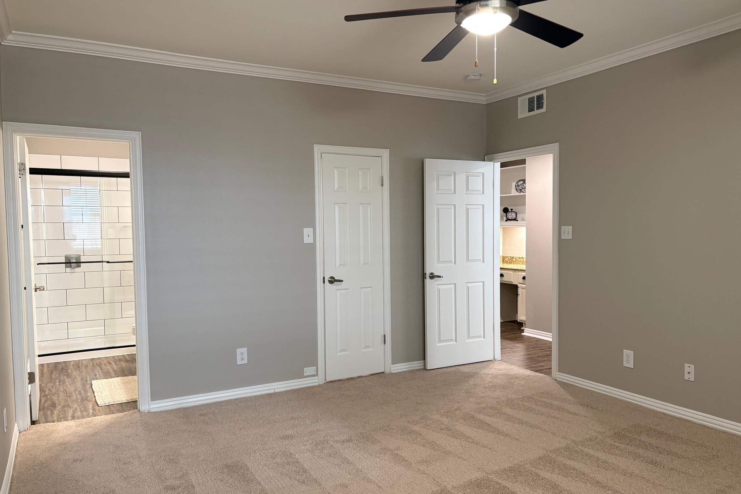 Empty room featuring beige carpet and light gray walls. There are three doors: one leading to a bathroom with a white tile shower visible through an open door, another leading to a small kitchen area, and the third closed. A ceiling fan hangs from the ceiling, providing a modern touch to the space.