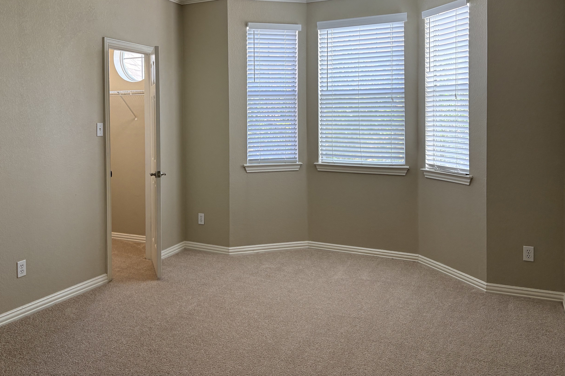 A vacant room with light beige walls and carpet. It features three windows with blinds, providing natural light. A door on the left leads to a small closet space. The room has a cozy, minimalist design, perfect for various uses.