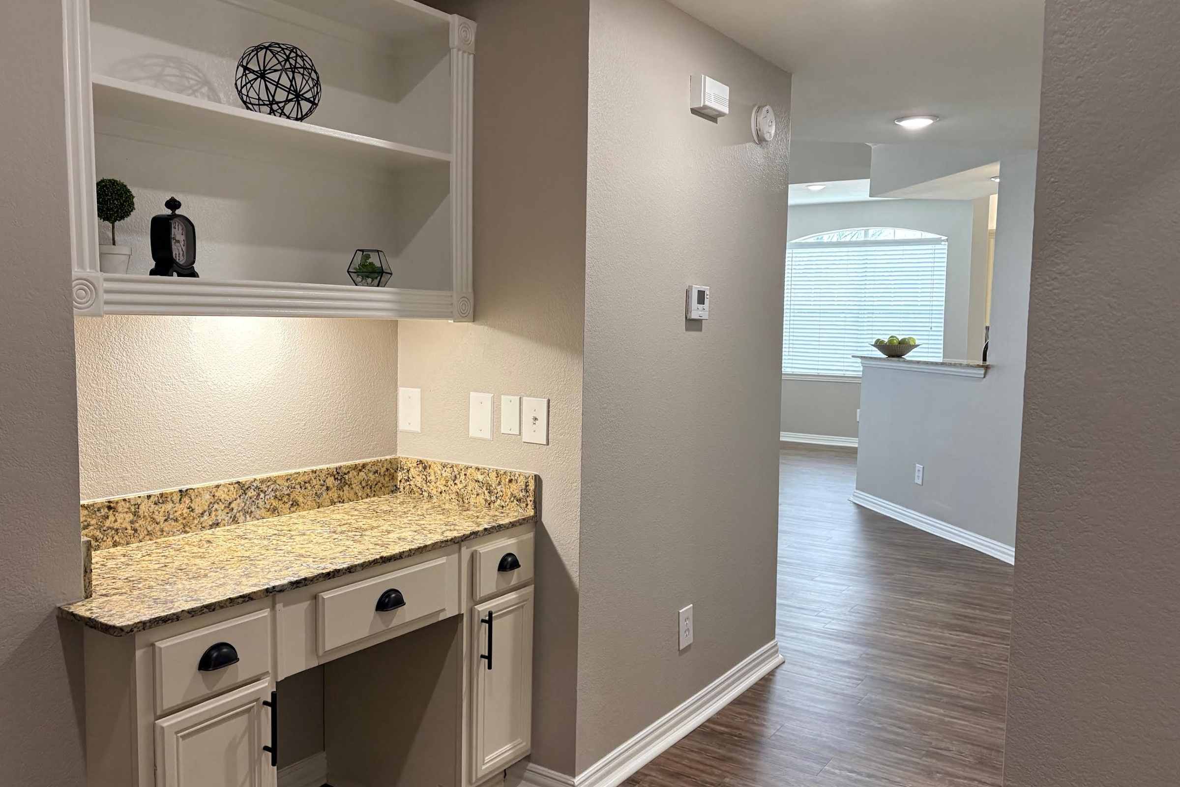 Interior view of a home featuring a hallway with granite countertop cabinetry, wall outlets, and decorative items on a shelf. The background shows a well-lit living area with a window and a neutral color scheme. The flooring is a warm wood tone, enhancing the spacious feel of the layout.
