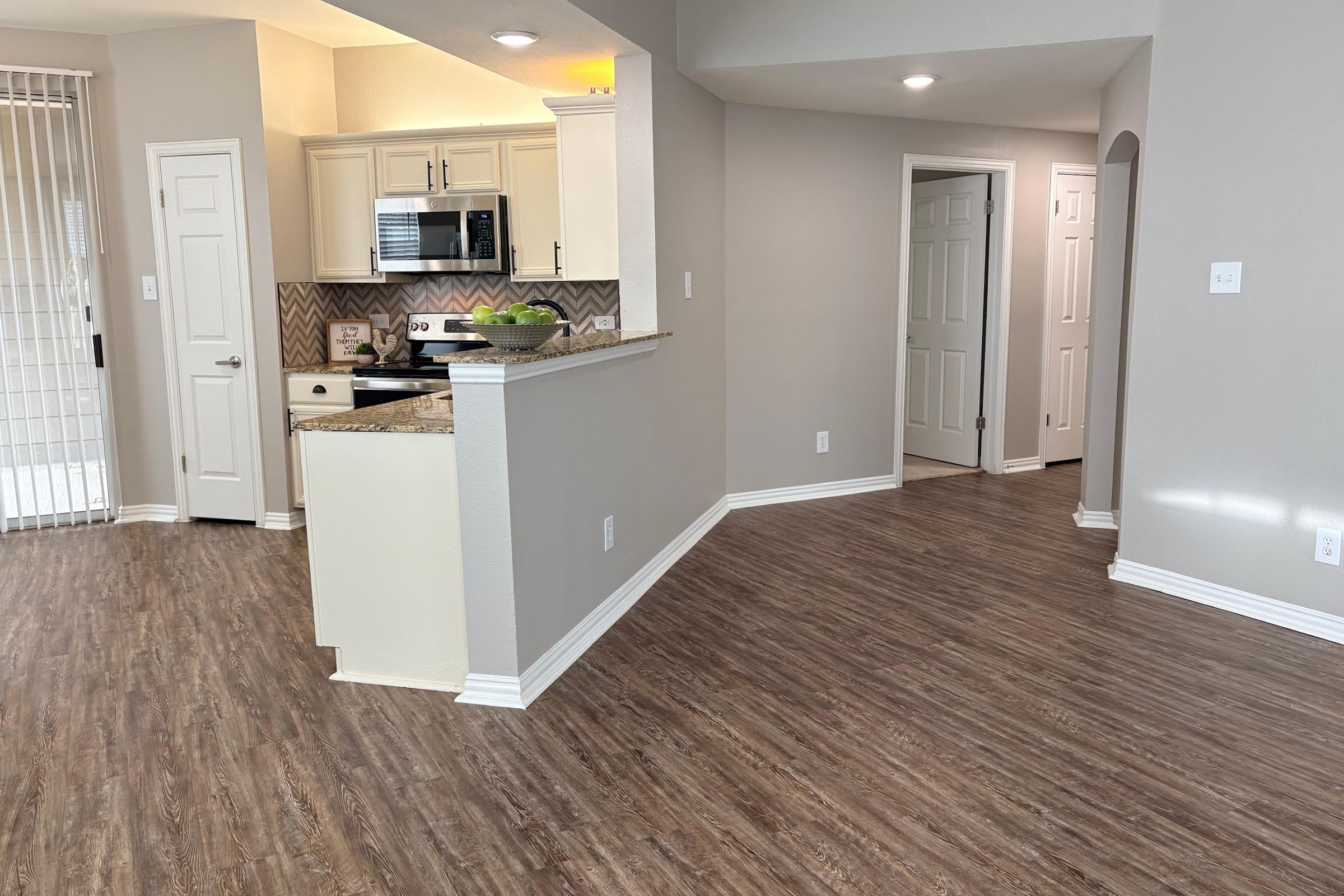 Interior view of a modern living space, featuring an open floor plan. The kitchen is partially visible with light cabinetry, a microwave, and granite countertops. A hallway leads to another room, while the floor has a wood-like finish. Bright natural light flows in from a window, enhancing the inviting atmosphere.