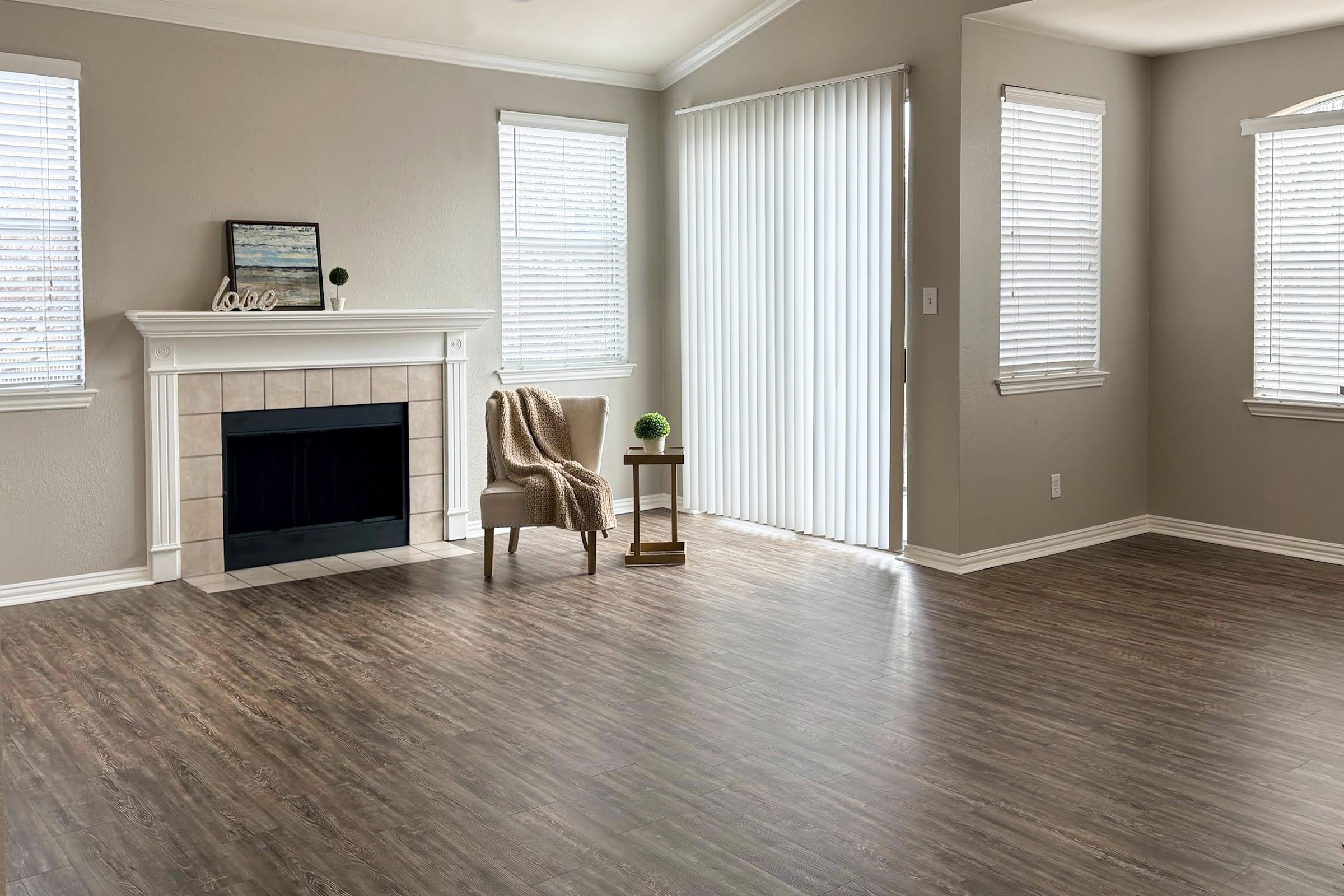 A well-lit, empty living room featuring a cozy beige armchair next to a small side table. There’s a fireplace with a simple decorative piece on the mantel. The room has large windows with white blinds, and the flooring is a warm, wood-like laminate, creating a spacious and inviting atmosphere.
