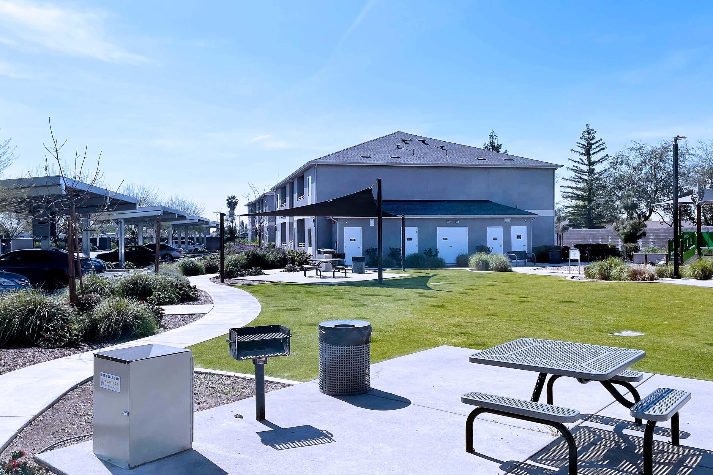 A spacious outdoor area featuring a green lawn, several picnic tables, and a trash bin. In the background, there is a two-story building with white doors and shaded parking spaces. Sparse trees and shrubs enhance the landscape, creating a pleasant environment for relaxation and social gatherings.