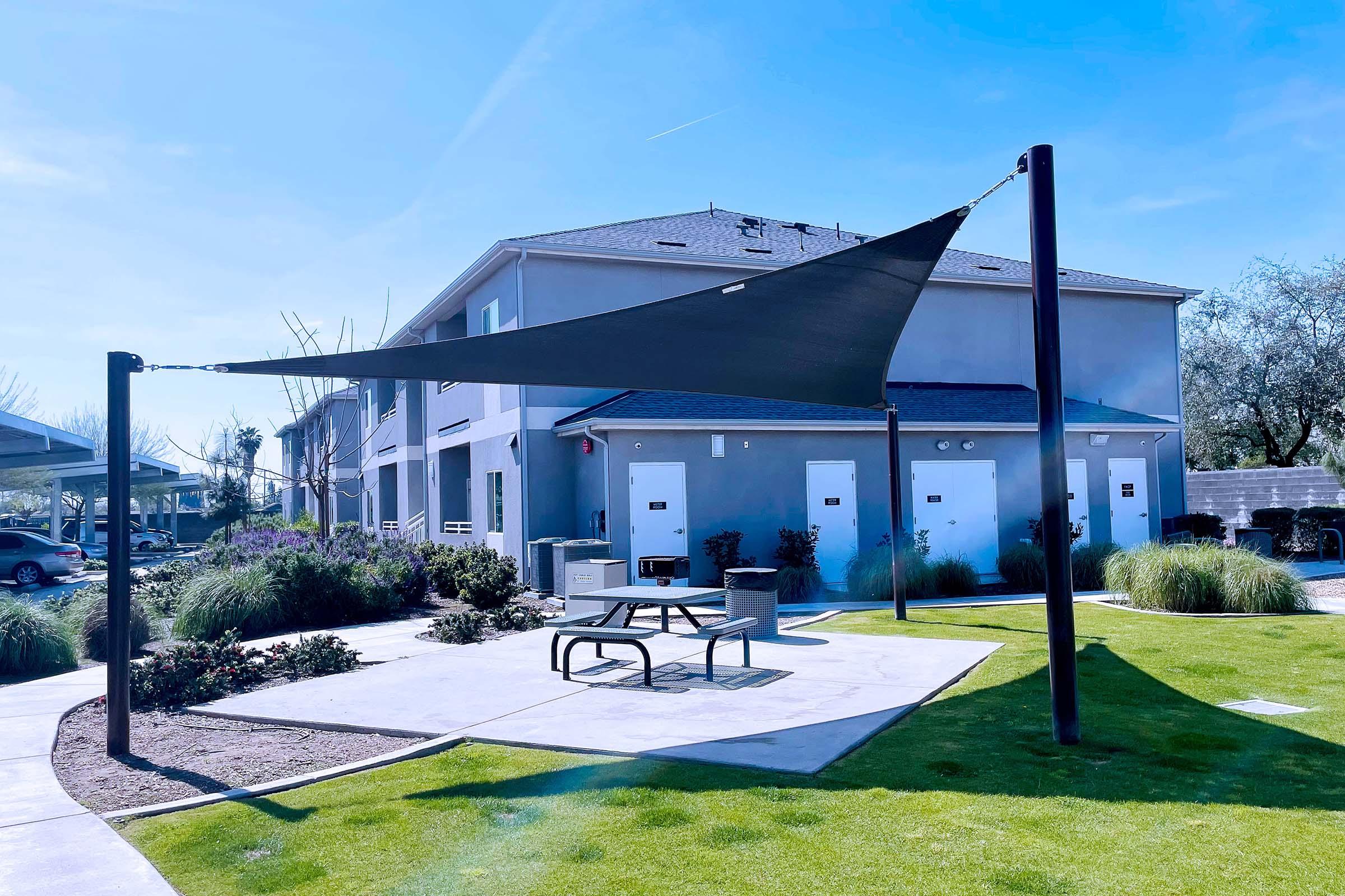 A triangular shade sail supported by two poles in a grassy area beside a modern building. There are picnic tables underneath the sail, and the surroundings include landscaped bushes and pathways. The sky is clear with some clouds.