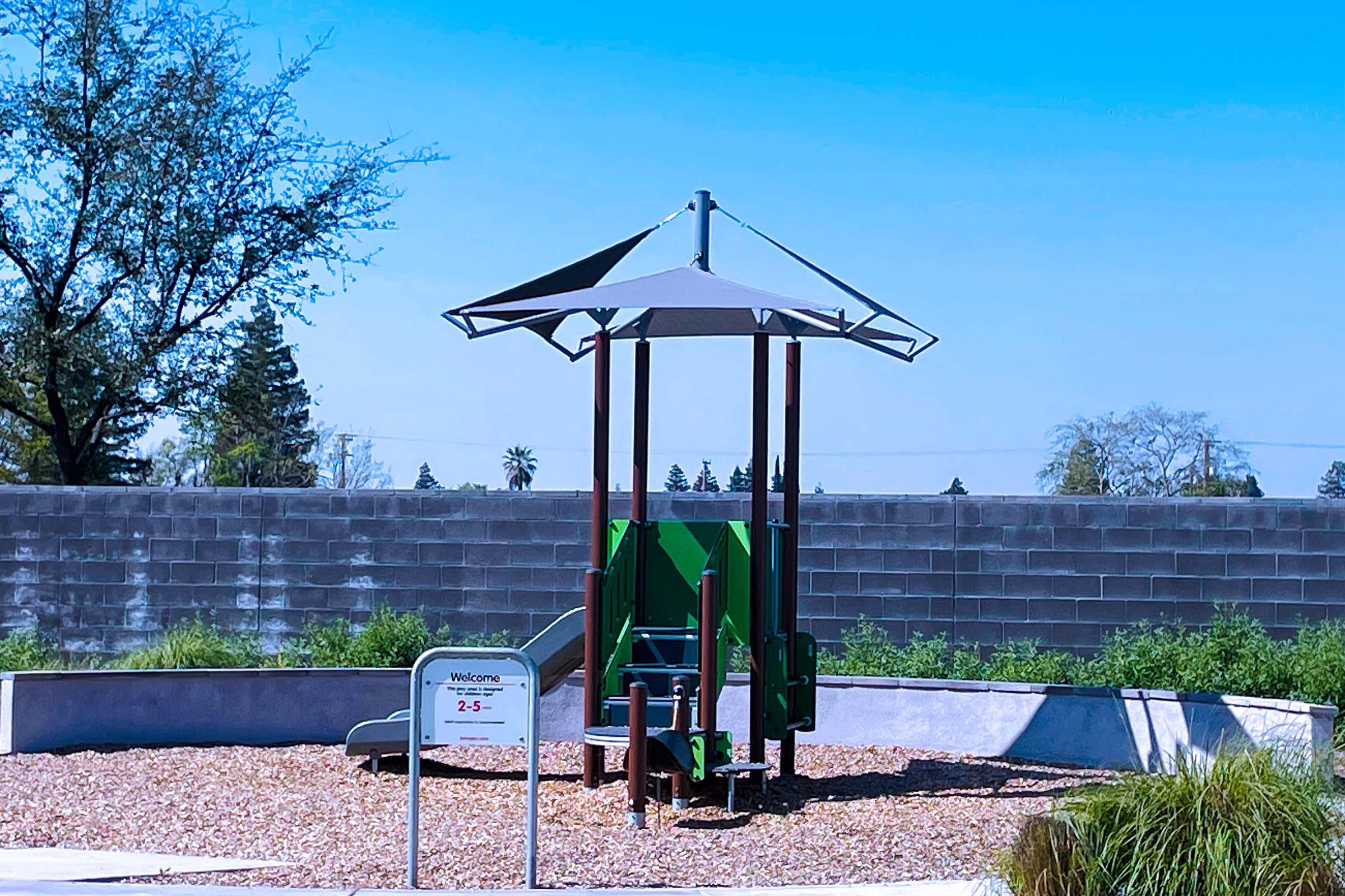 A playground featuring a green play structure with a shaded canopy, surrounded by gravel and green grass. There is a sign welcoming visitors, and a gray wall in the background with trees and clear blue skies above.