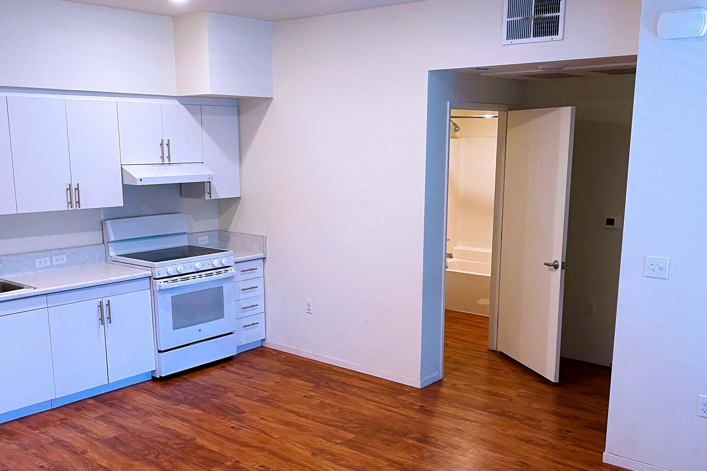 Modern kitchen with white cabinets and a stove, featuring a clean countertop. There's a doorway leading to a bathroom on the right, which has a light-colored interior. The floor is made of brown wood, and the walls are painted a soft beige. Overall, the space appears bright and inviting.