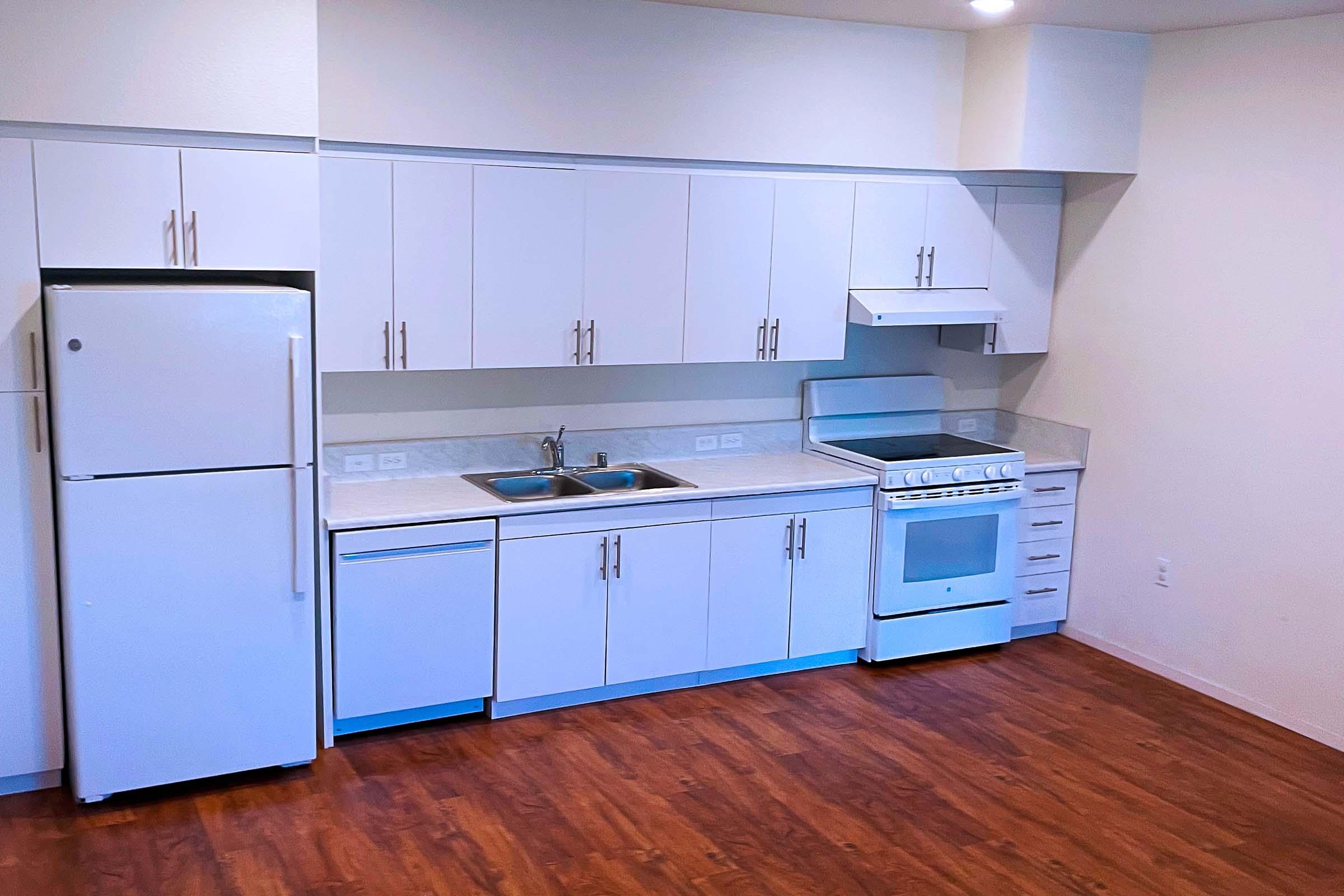 A clean, modern kitchen featuring white cabinets, a stainless steel refrigerator, a gas stove, and a double sink. The countertops are light-colored, and the flooring is wood. The kitchen has a minimalist design, with ample cabinet space and a bright, airy feel.