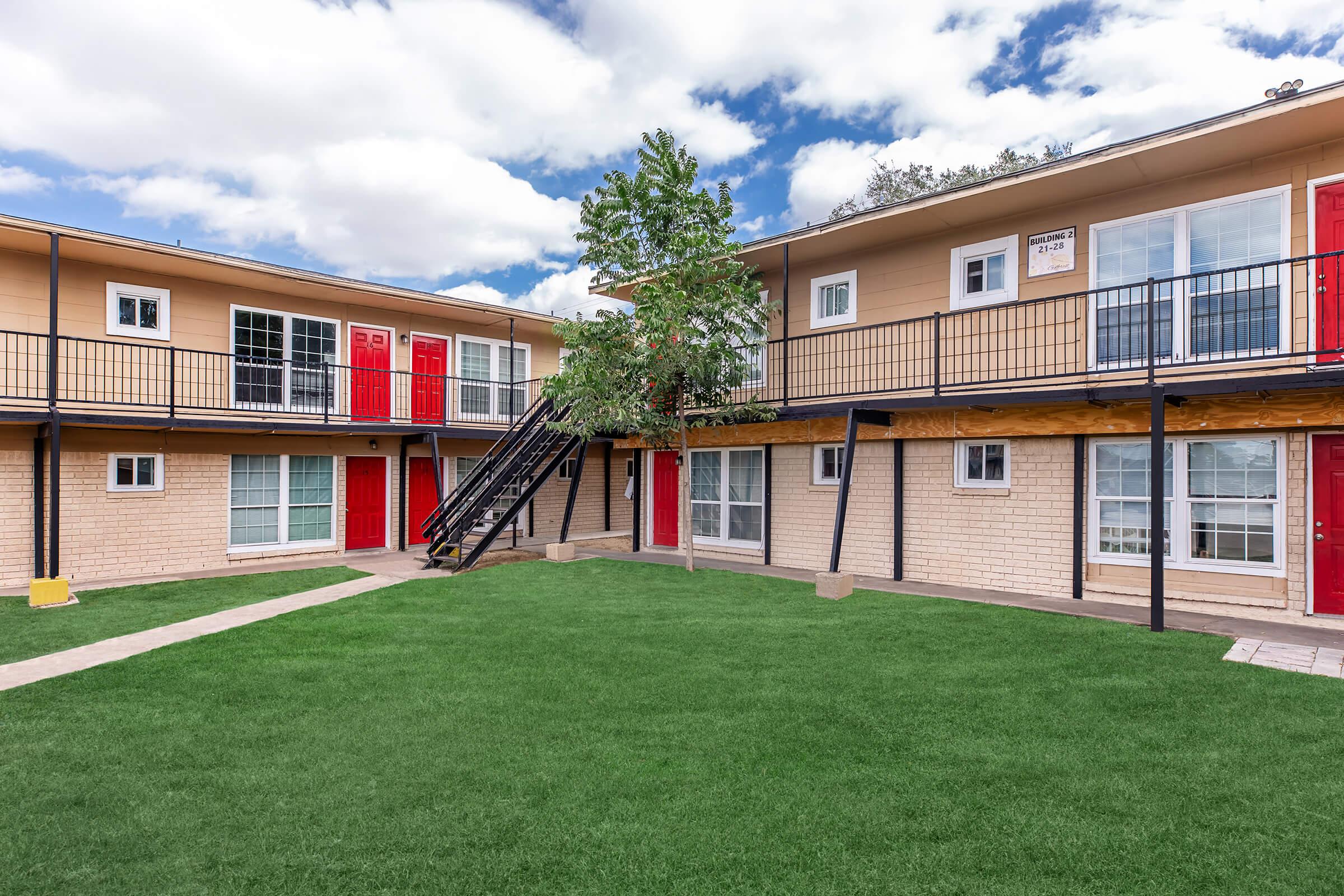 A view of a two-story apartment complex featuring several units with red doors and balconies. There is a grassy courtyard in the center, with a walkway leading through it. The sky is partly cloudy, adding natural light to the scene.