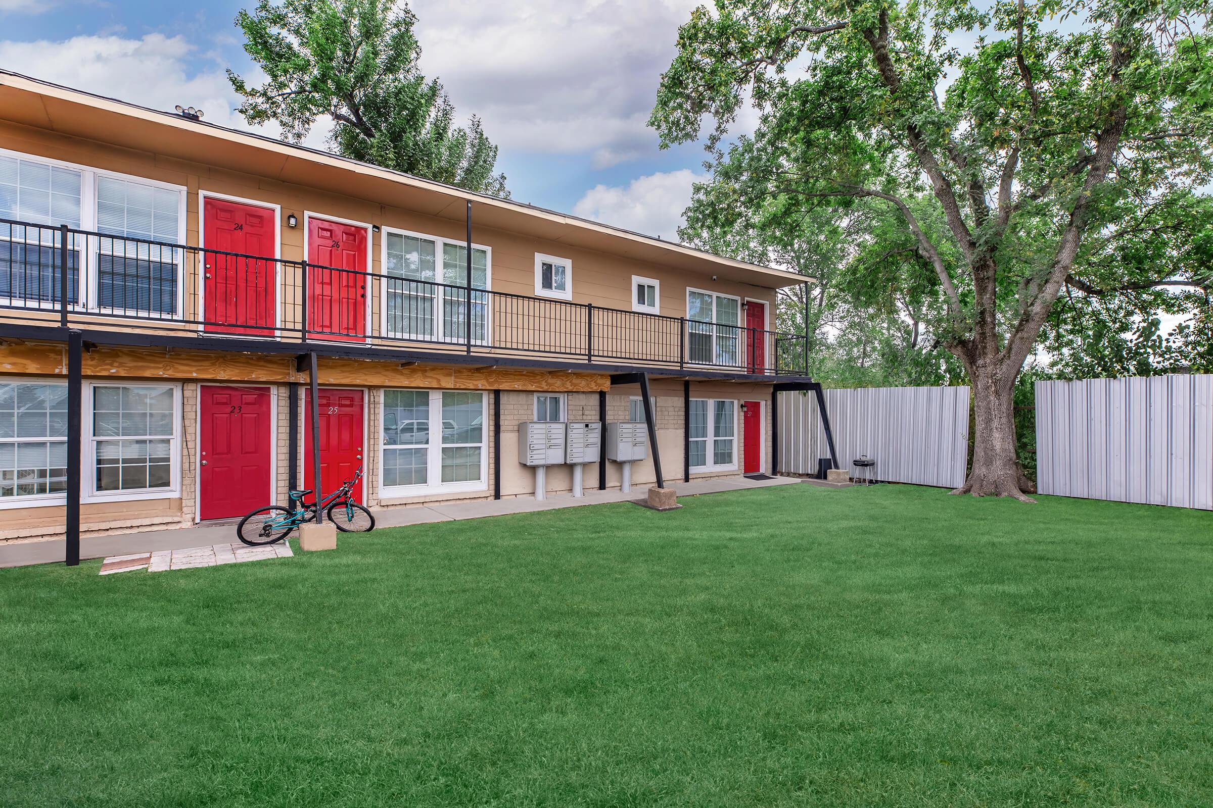 Two-story building with a beige exterior featuring red doors on both levels. A small green lawn is in front with a bicycle parked on the left side. The surrounding trees are lush, and a white fence is visible in the background, with a cloudy sky overhead.