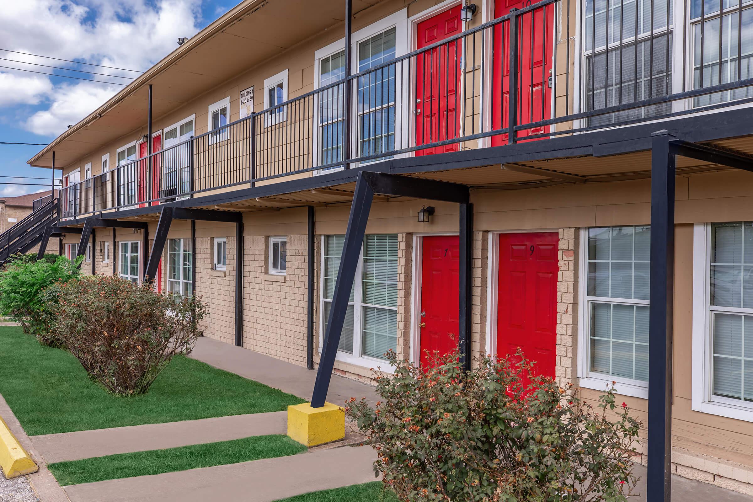 A view of a two-story motel with a beige exterior and red doors. The building features a balcony with black railings, surrounded by neatly trimmed bushes and small patches of grass. The sky is partly cloudy, adding a bright atmosphere to the setting.
