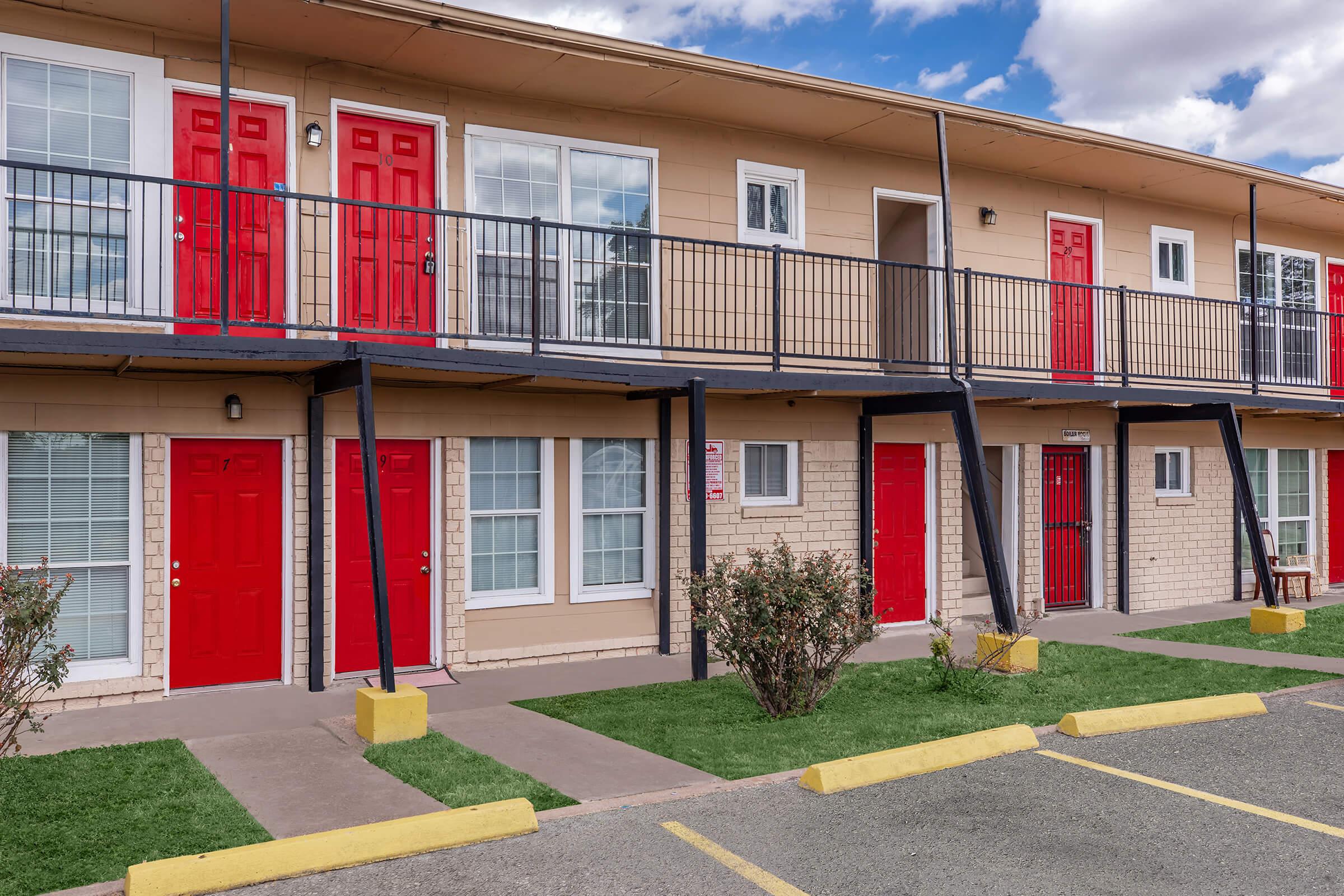 A row of two-story apartment units with beige walls and red doors, featuring balconies on the upper level. Green grass borders a concrete pathway, and parking spaces are visible in the foreground. The sky above is blue with scattered clouds.