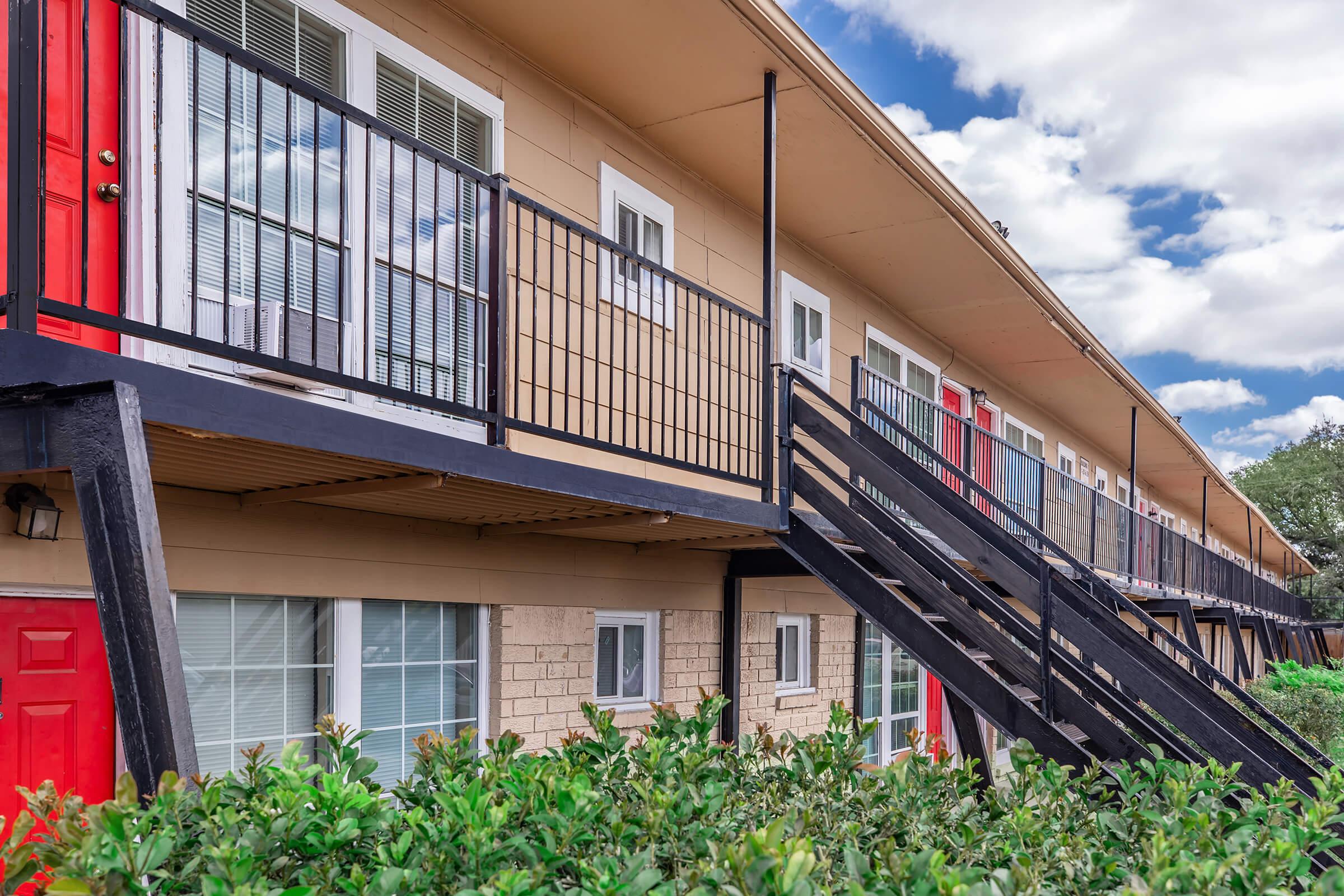 Two-story building with a balcony and exterior stairs. The building features multiple doors, some painted red, and large windows. A green hedge is visible at the base, with a partly cloudy sky in the background.