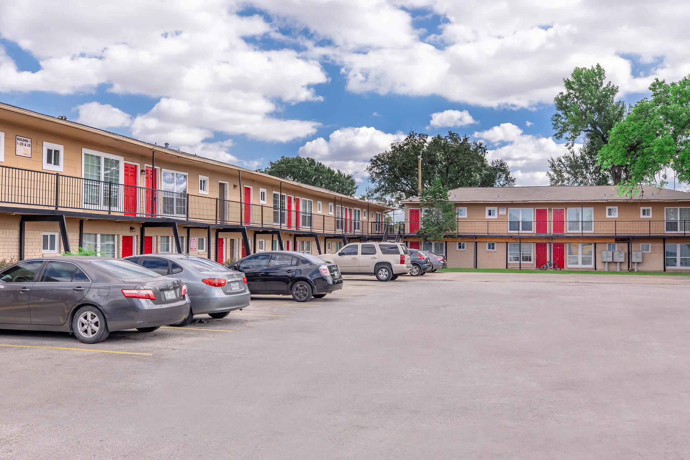 A view of a multi-unit apartment complex featuring two-story buildings with red and beige doors. Several parked cars occupy the foreground, and lush green trees can be seen in the background under a partly cloudy sky.