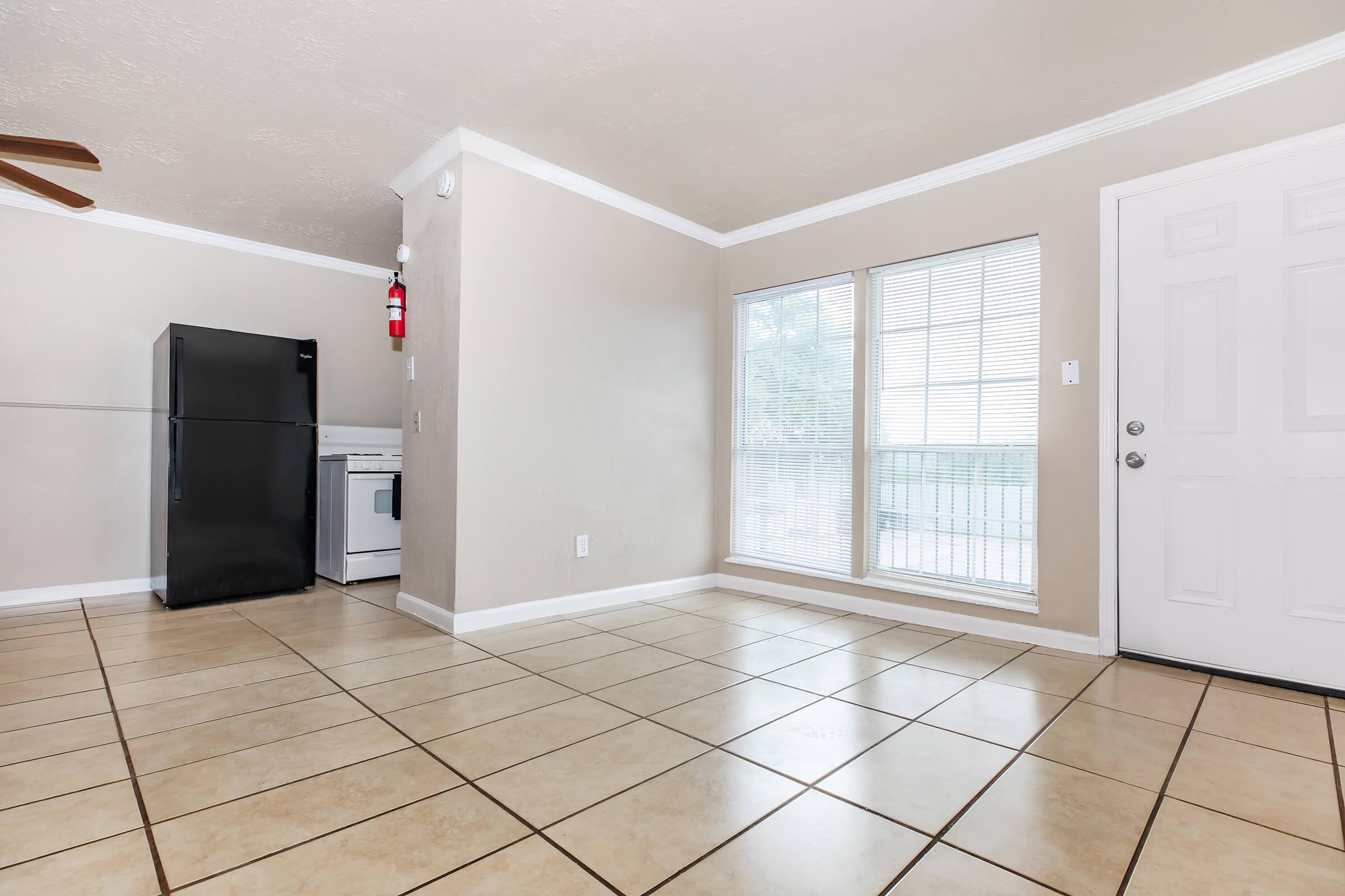 Empty living space featuring a tiled floor, a black refrigerator, and a white washing machine. There is a large window with blinds allowing natural light, and a door leading outside. The walls are painted in a neutral color, and a ceiling fan is visible.