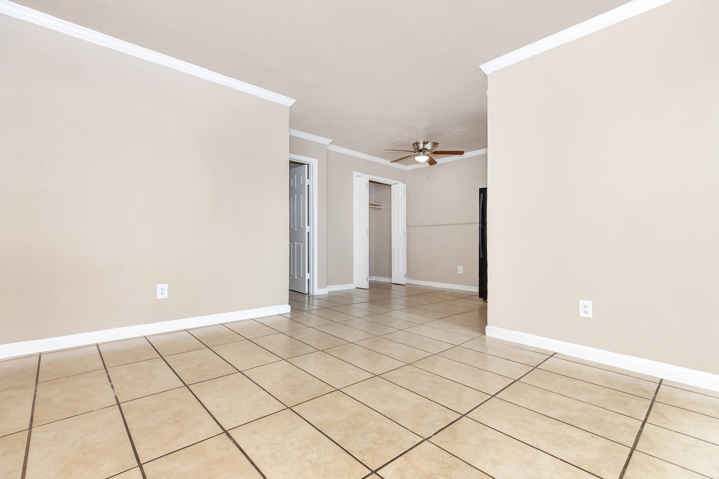 Spacious interior of a room with beige walls and tiled flooring. A ceiling fan is visible, along with doorways leading to other areas. The room is well-lit and uncluttered, offering a blank canvas for potential furnishings or decor.