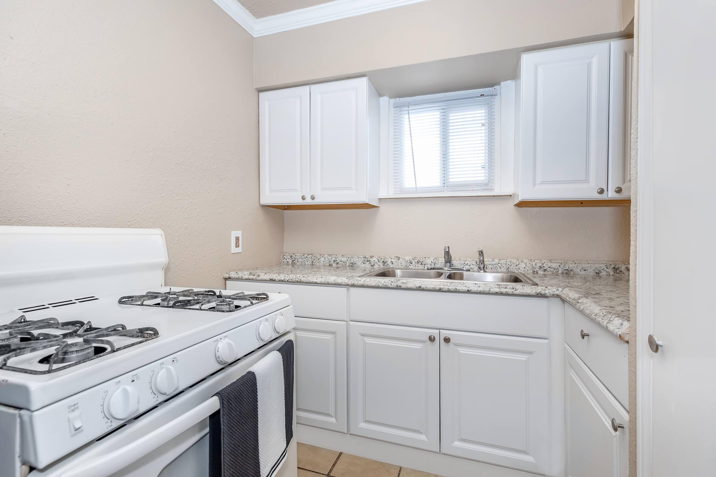 Compact kitchen featuring white cabinetry, a granite countertop, a gas stove, and a double sink under a window. The walls are painted in a neutral tone, and the floor is tiled, creating a clean and functional cooking space.