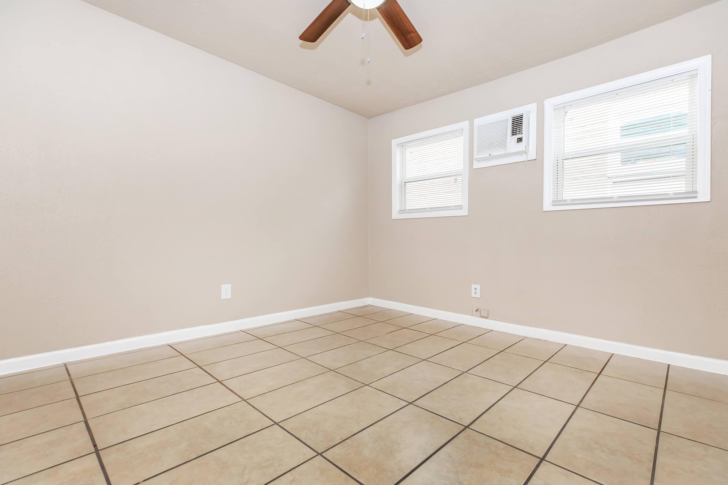 A small, empty room with beige walls and tile flooring. There are two windows with blinds, an air conditioning unit, and a ceiling fan. The overall space appears clean and well-lit, suitable for various uses.