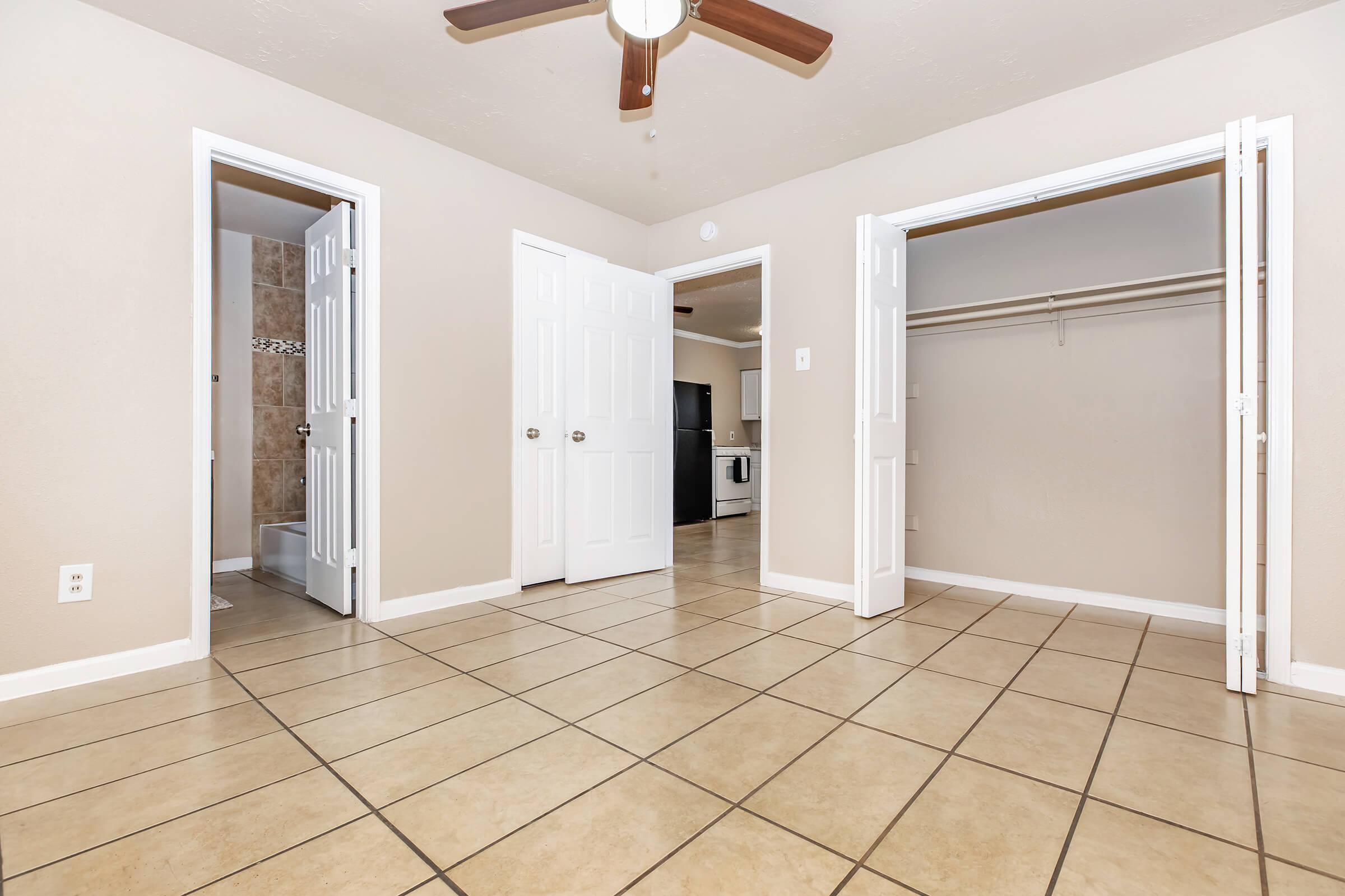 A spacious interior view of a room featuring tiled flooring. Two open doors lead to adjoining areas, including a closet and a bathroom. In the background, a doorway connects to a kitchen with visible appliances. The walls are painted light colors, and a ceiling fan is located above.