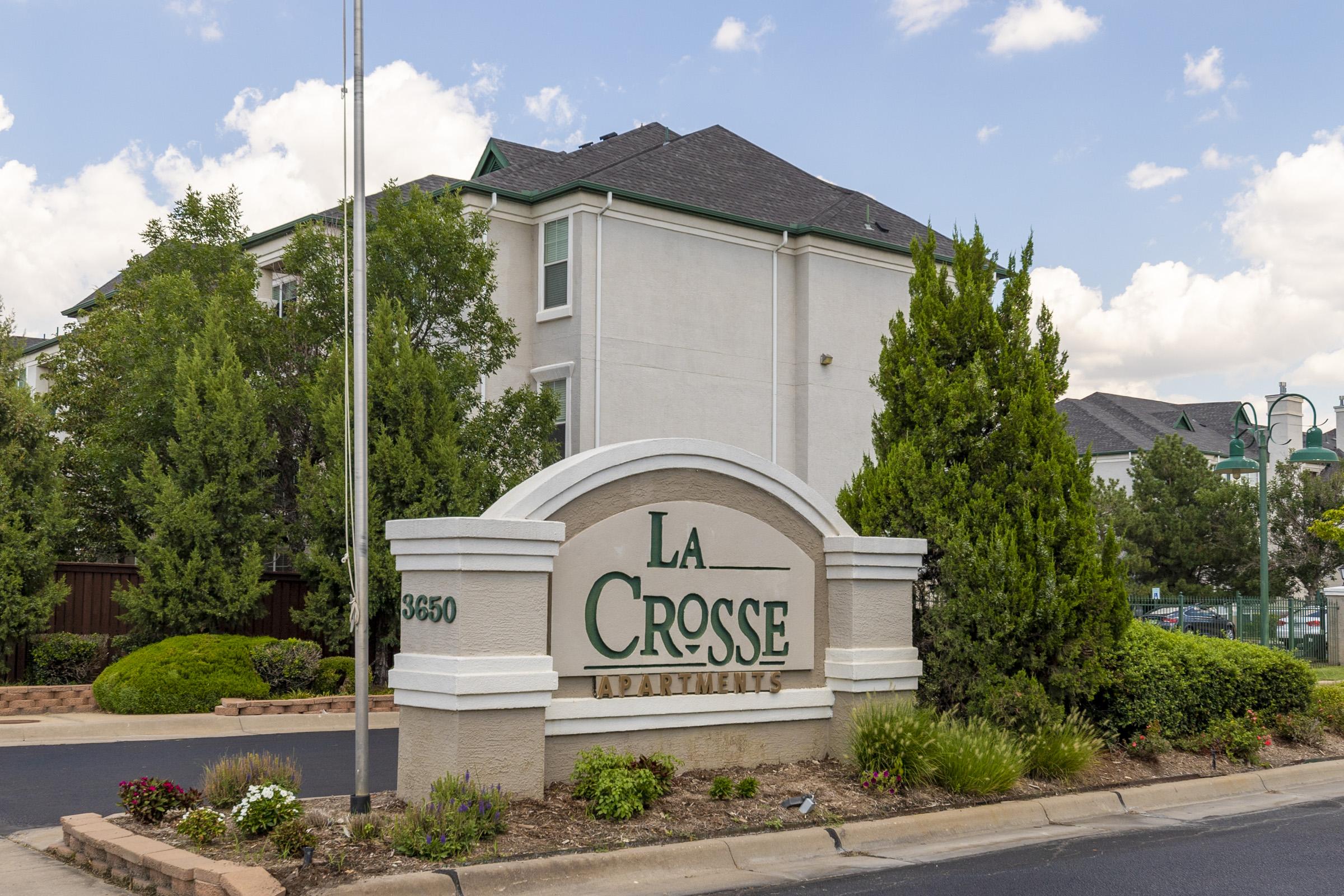 Sign for La Crosse Apartments, displaying the name prominently. The background features a well-maintained exterior of an apartment building with greenery and landscaping. A clear blue sky with a few clouds adds to the image's ambiance.
