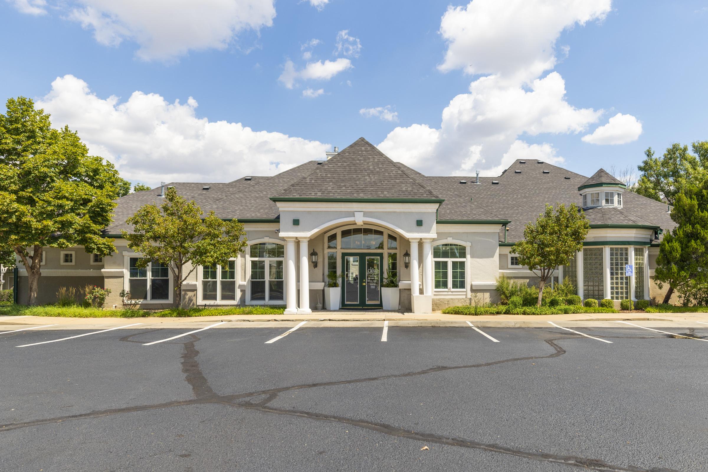 A light-colored building with a peaked roof and a portico entrance, surrounded by greenery. The parking lot in front has several empty spaces, and the sky above is partly cloudy. The architectural style is modern with decorative elements.