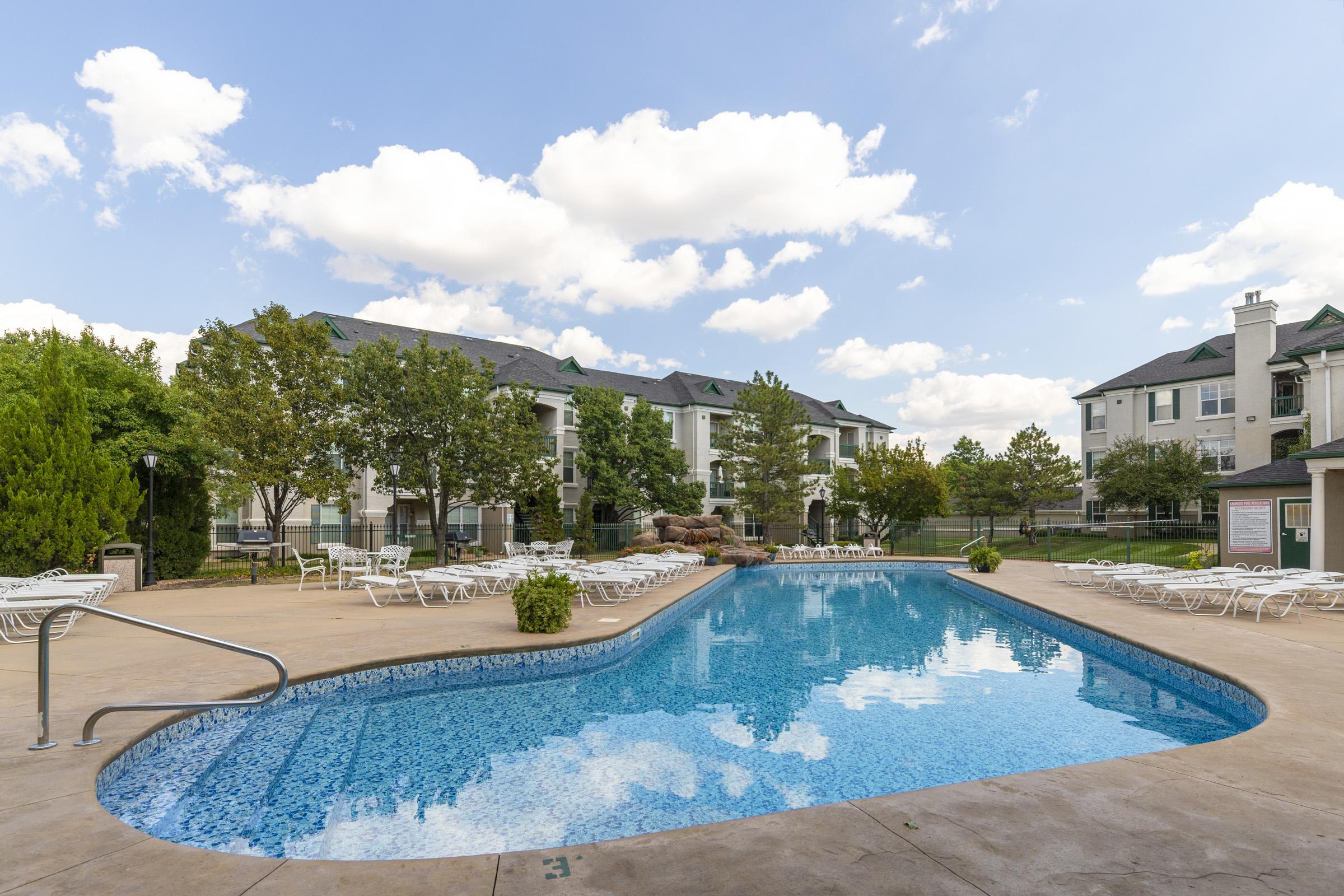 A serene outdoor pool area surrounded by lounge chairs and trees, with modern apartment buildings in the background. The pool's clear blue water reflects the sky with fluffy white clouds. Ideal for relaxation and leisure activities.