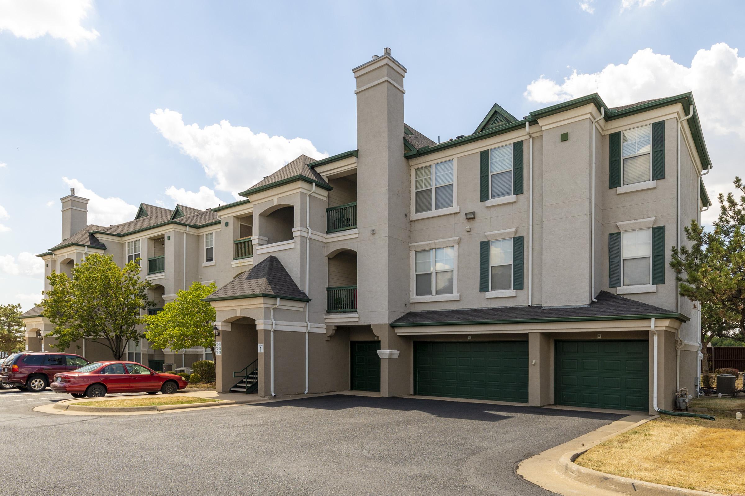 A three-story apartment building with a light beige exterior, green accents, and balconies. The building features garages on the ground level. In the foreground, there are parked cars, including a red vehicle and surrounding greenery. The sky is partly cloudy.