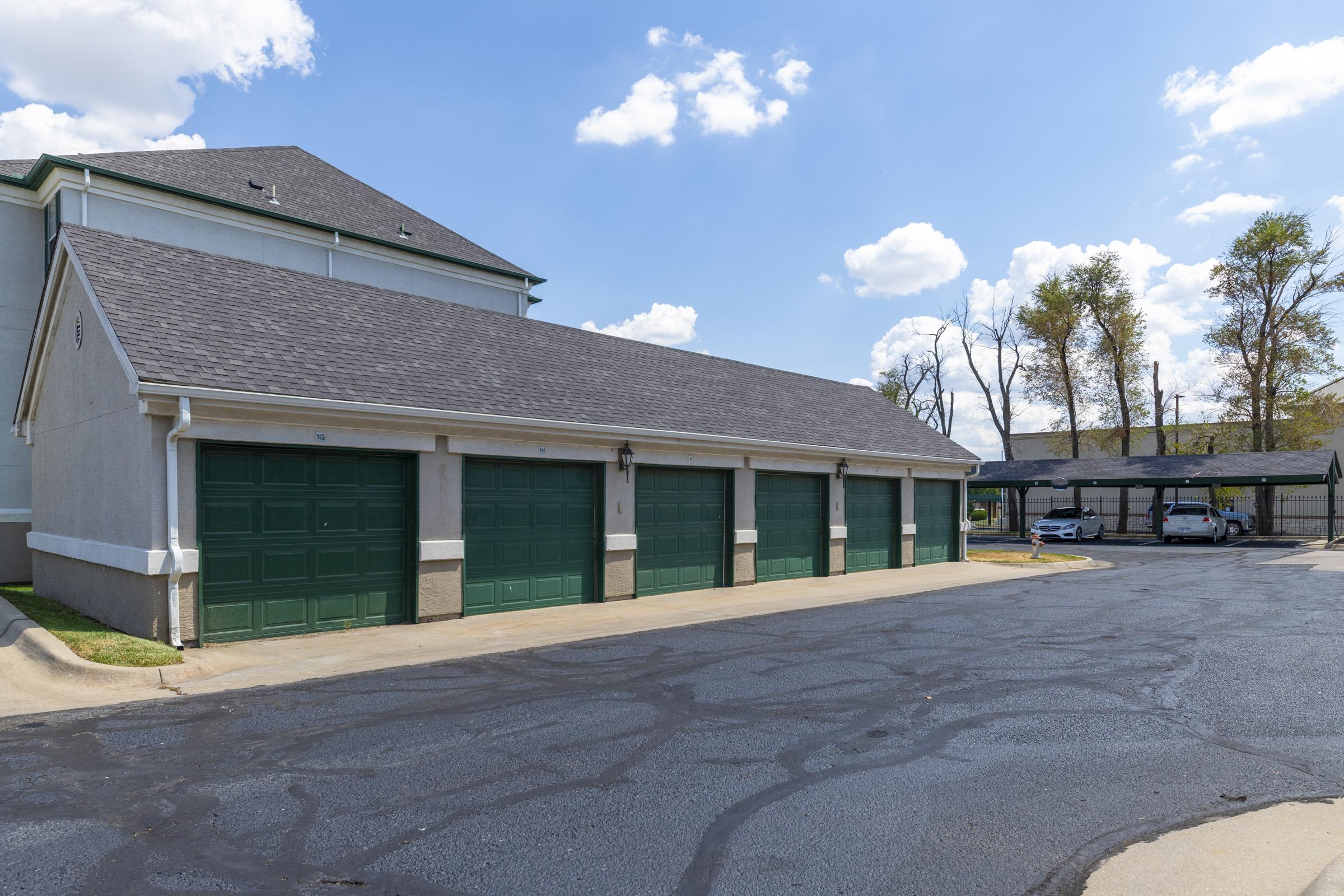 A row of green garage doors against a light-colored building, with a newly paved black asphalt driveway in the foreground. The sky is blue with scattered clouds, and there are trees in the background, indicating a suburban setting.