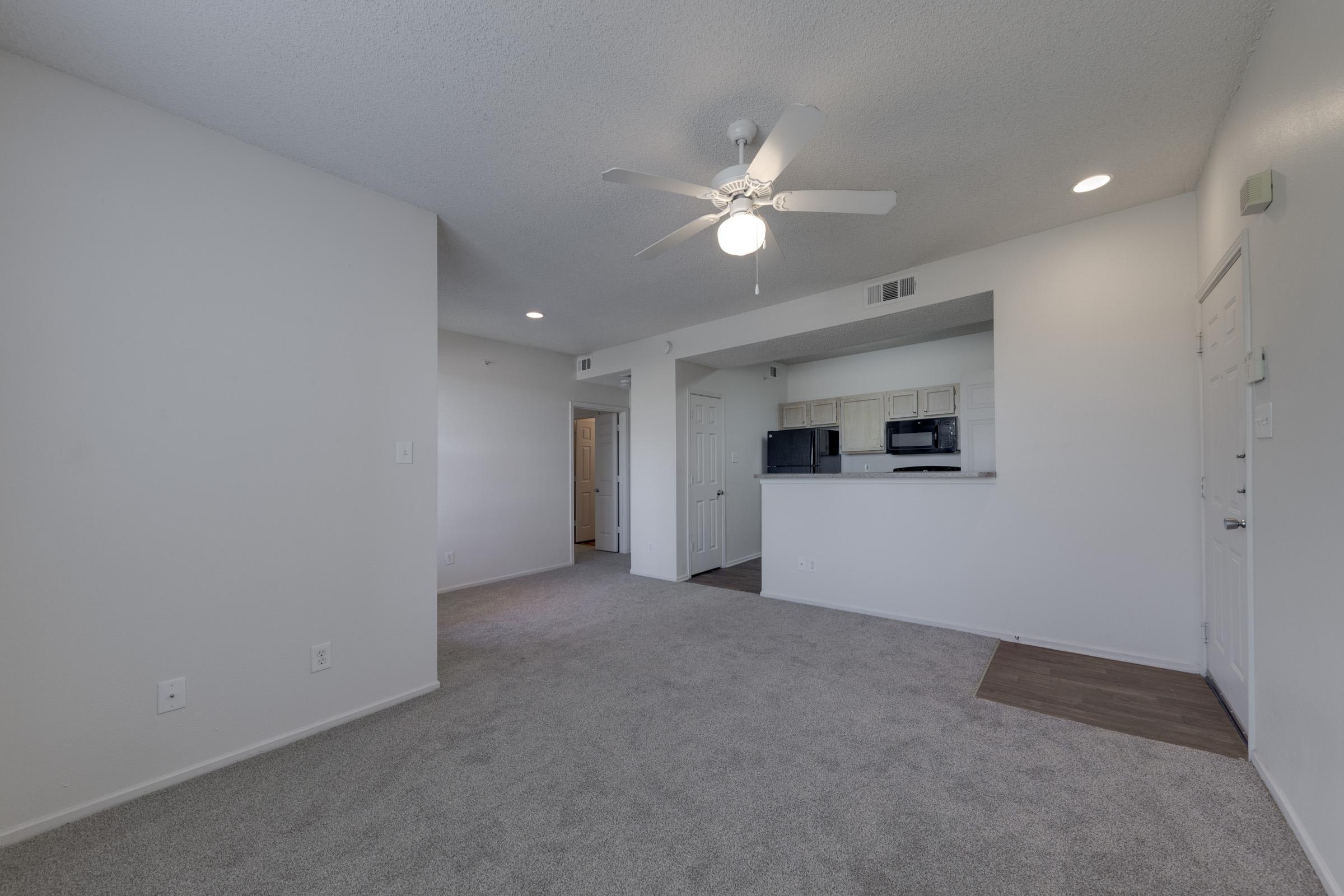 A spacious, well-lit room with beige carpet, featuring a ceiling fan, and an open layout leading to a kitchen area with light cabinets. A doorway can be seen in the background, suggesting additional rooms. The walls are painted white, providing a clean and neutral environment.