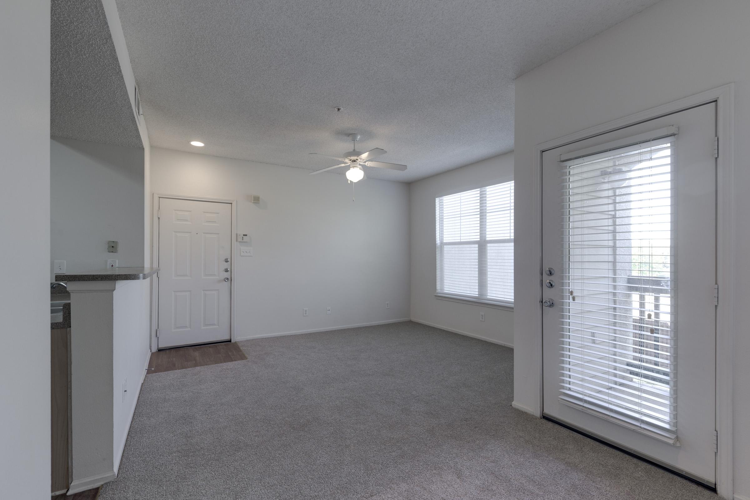 A spacious, empty room with light-colored walls and carpet. It features a ceiling fan, large window with blinds, and a sliding glass door leading outside. The entrance area includes a front door and a small wall section with a counter space nearby. Ideal for a living area or a den.
