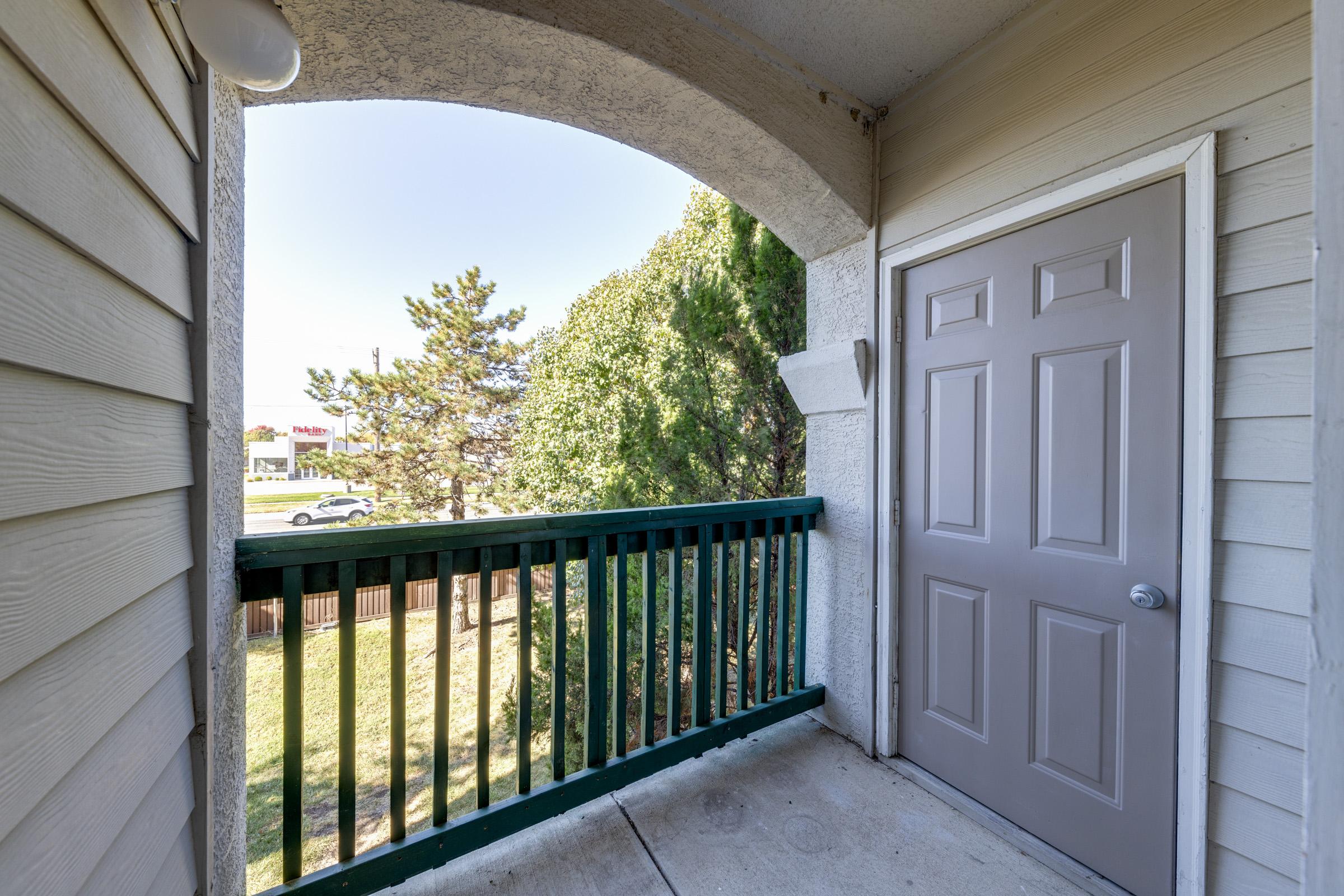 A small outdoor balcony with a green railing, featuring a view of trees and a road in the distance. The entrance door is grey, and the walls are light-colored wood. The space is well-lit by natural sunlight filtering in from outside.