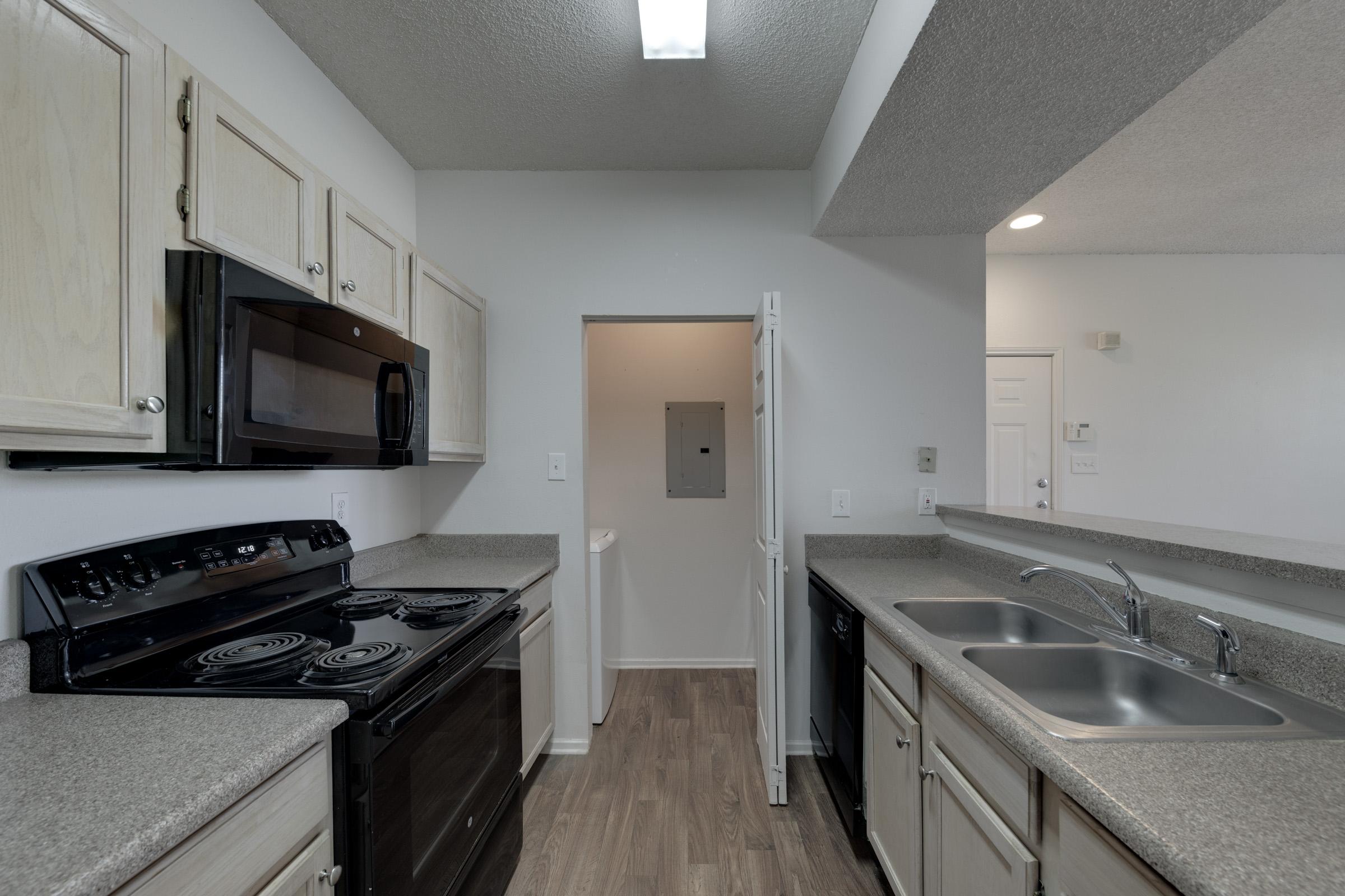 A modern kitchen featuring light wooden cabinetry, a black microwave above a black stove, and dual stainless steel sinks. The countertops are gray with a textured finish, and the flooring is light wood. A doorway leads to another room in the background, and there is overhead lighting in the ceiling.