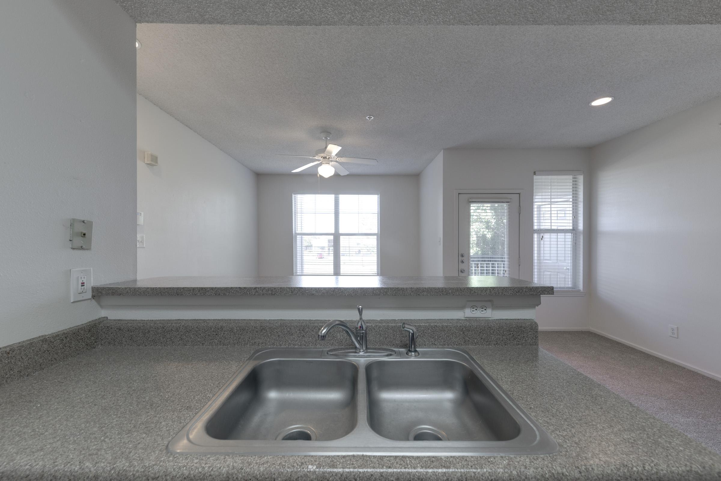 A modern kitchen view with a double sink and granite countertop, featuring a light ceiling fan above. In the background, there is a spacious living area with large windows allowing natural light in, and a door leading outside. The walls and carpets are neutral-colored, creating a bright, inviting atmosphere.