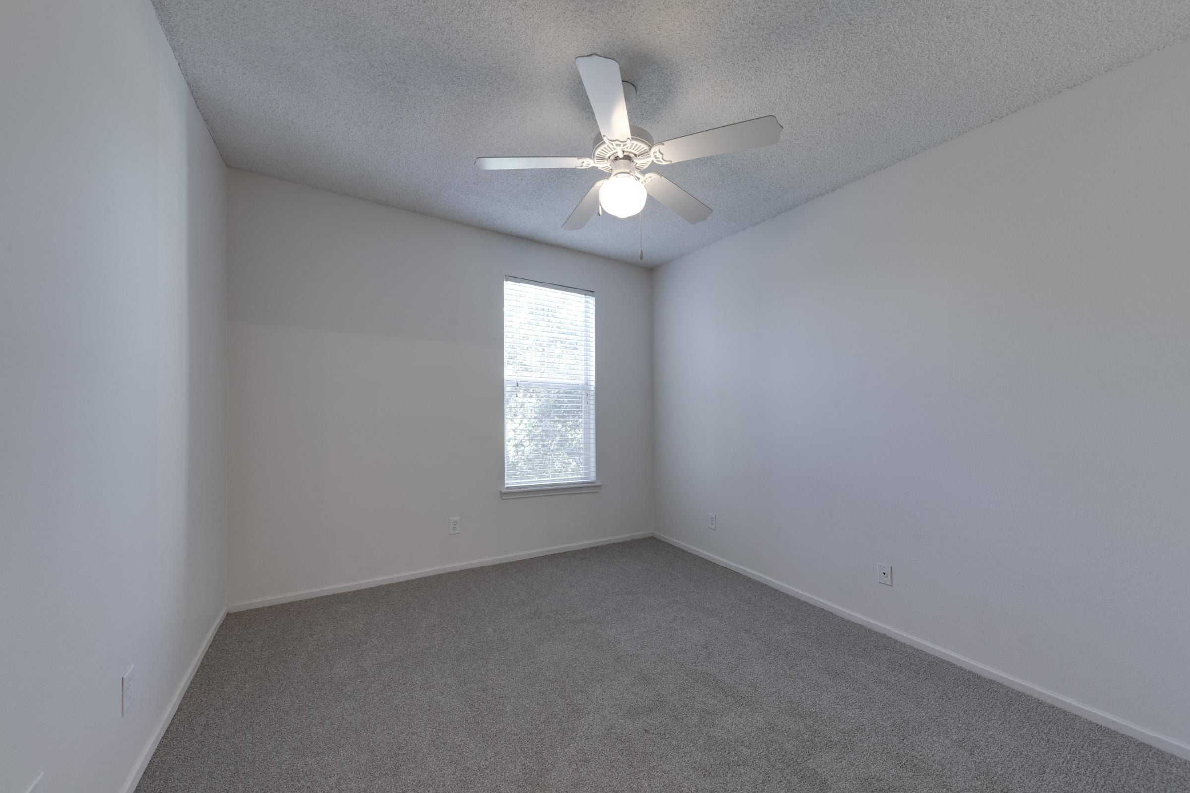 A spacious, empty room with light gray carpet, a ceiling fan with blades, and a window allowing natural light to illuminate the neutral-colored walls. The room has no furniture, creating a clean, open space ready for customization.