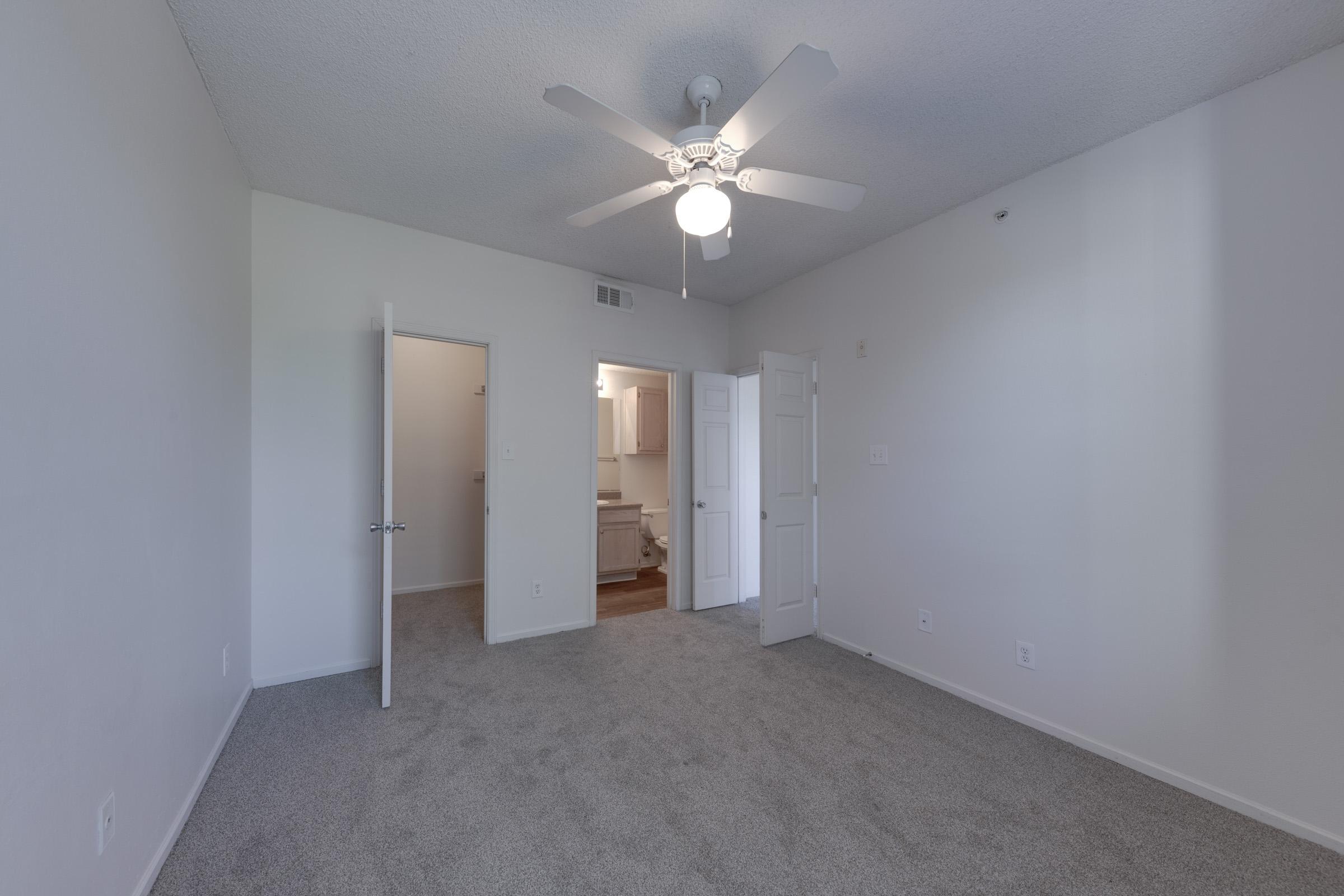 A well-lit, empty room with gray carpet and white walls. A ceiling fan is visible, and there are two doors: one leading to a bathroom and another to a hallway. The room is spacious, with plenty of natural light and minimal decor, creating a clean and welcoming atmosphere.