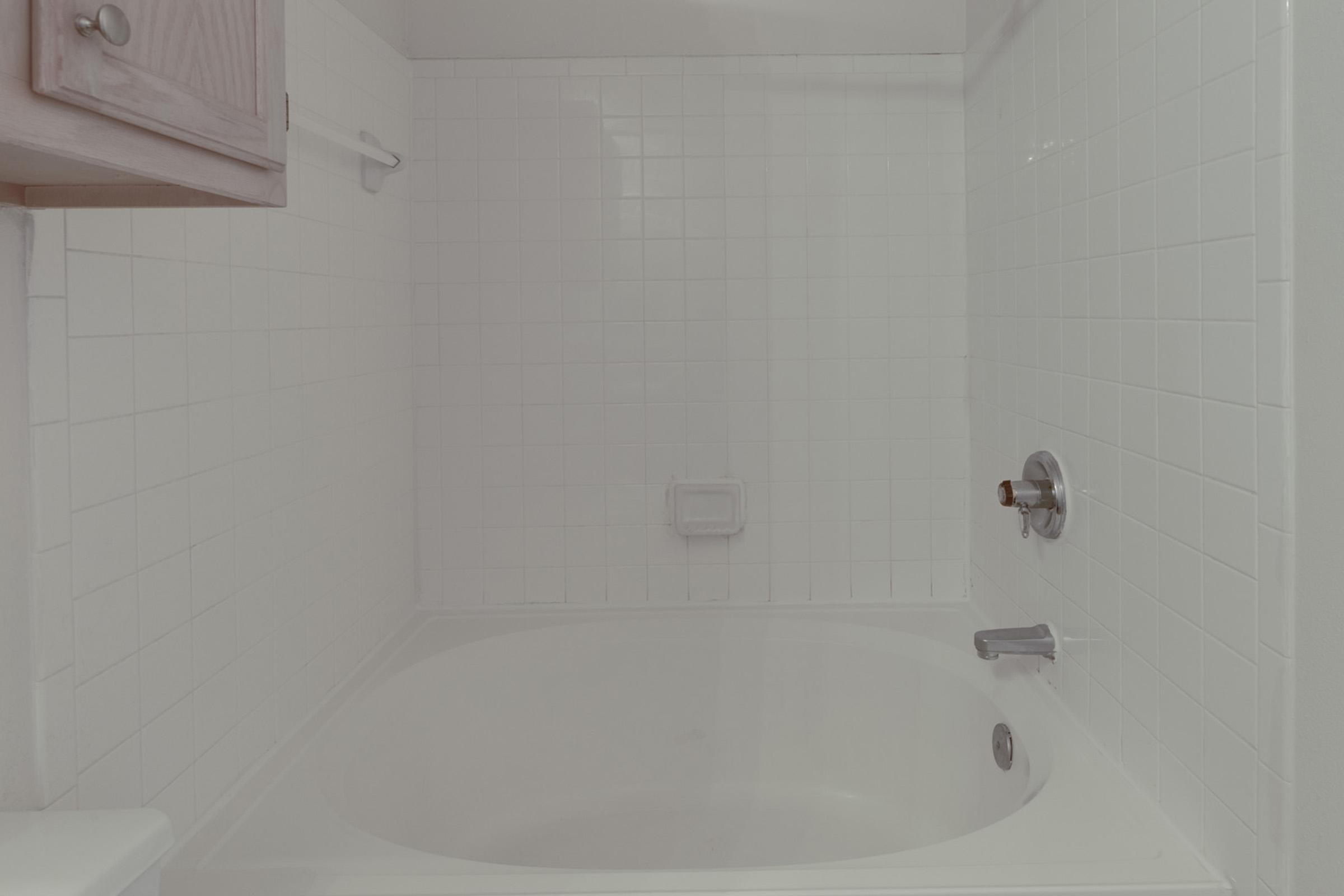 A clean, white bathroom with a bathtub and tiled walls. The bathtub is empty, and there are no visible toiletries or decorations. A wooden cabinet is partially visible on the left side. The overall aesthetic is minimalist and bright.