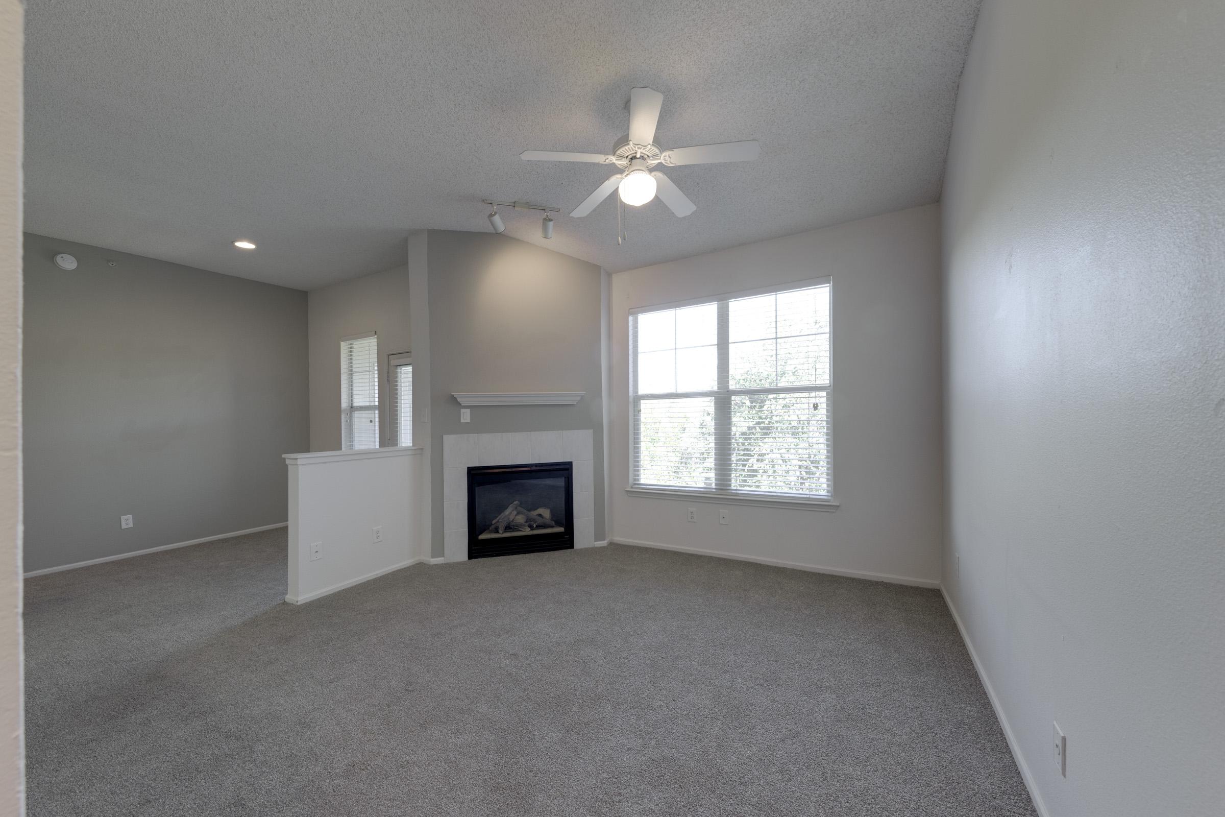 A spacious living room with light gray walls and carpet, featuring a ceiling fan and a small fireplace. Large windows allow natural light to fill the area, creating a bright and airy atmosphere. The room has a modern feel with minimal decor and an open layout leading to adjacent spaces.