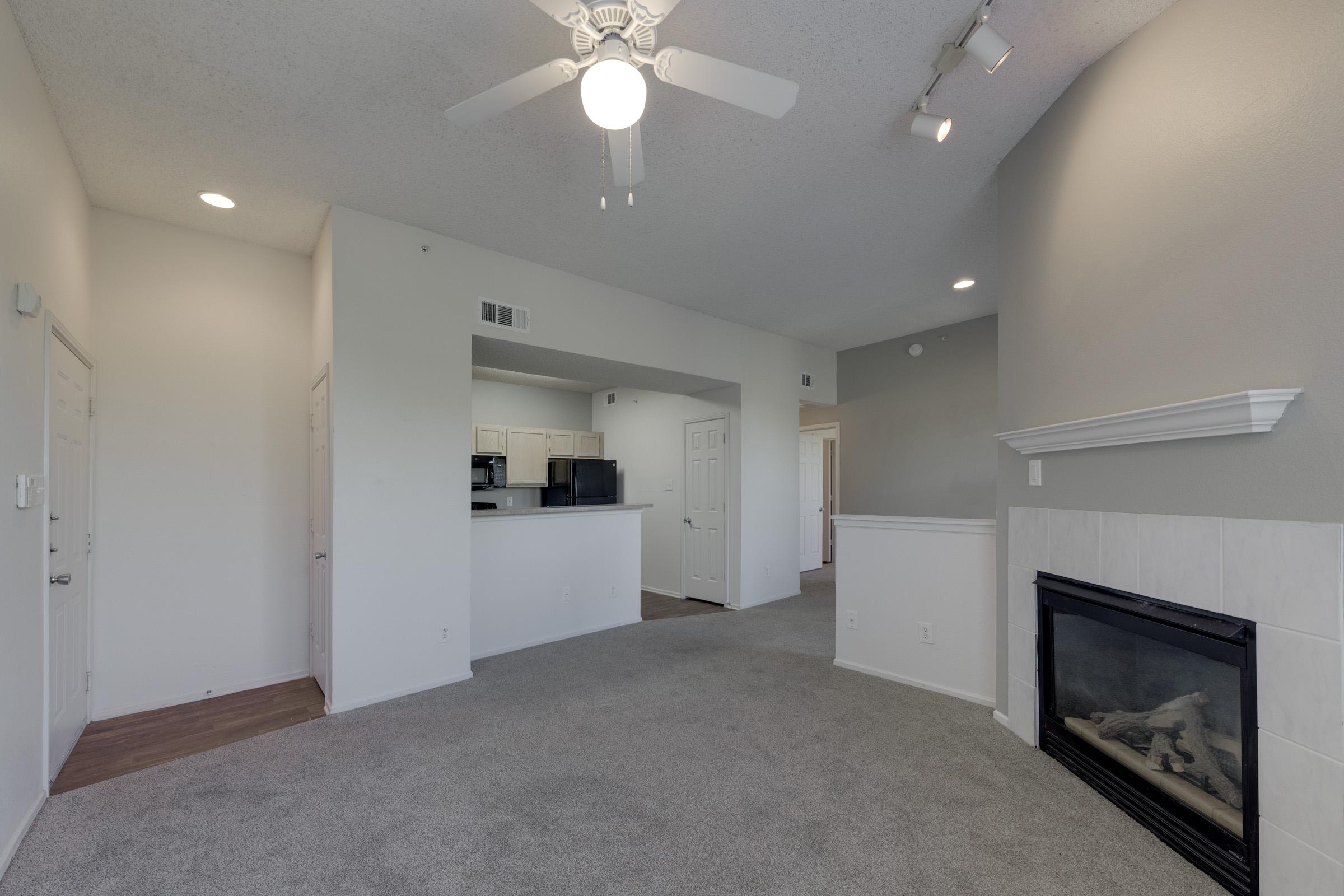 Interior shot of a contemporary living space featuring a light gray carpet, a ceiling fan, and a fireplace. The open layout connects to a kitchen area with white cabinetry and appliances visible in the background. Soft lighting from recessed fixtures enhances the inviting atmosphere.