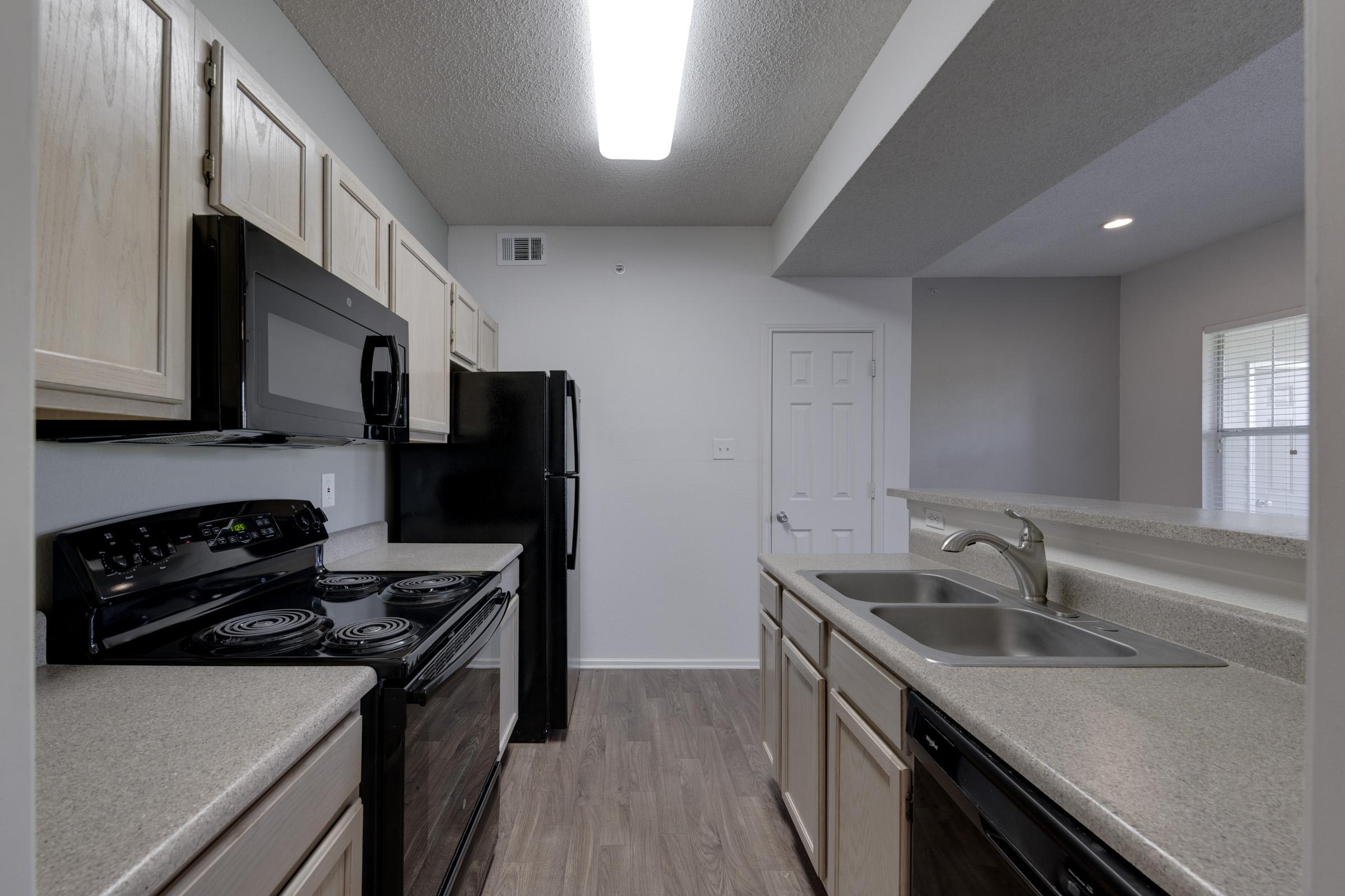 A modern kitchen featuring light wooden cabinets, a black microwave, stove, and refrigerator. The countertops are light-colored with a double sink. Natural light comes through a window, illuminating the space. The flooring is a light wood laminate.