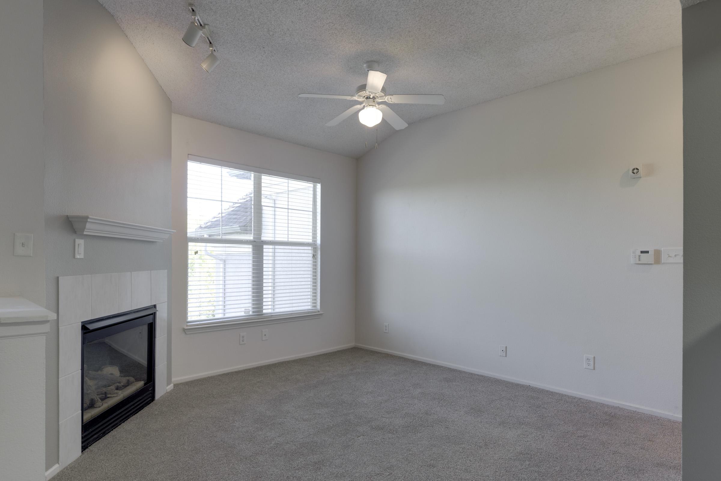 A bright, empty living room featuring a ceiling fan, a white fireplace, and a large window with blinds. The walls are painted in a light color, and the flooring is covered with light gray carpet. The overall space feels airy and open, ideal for personal customization.