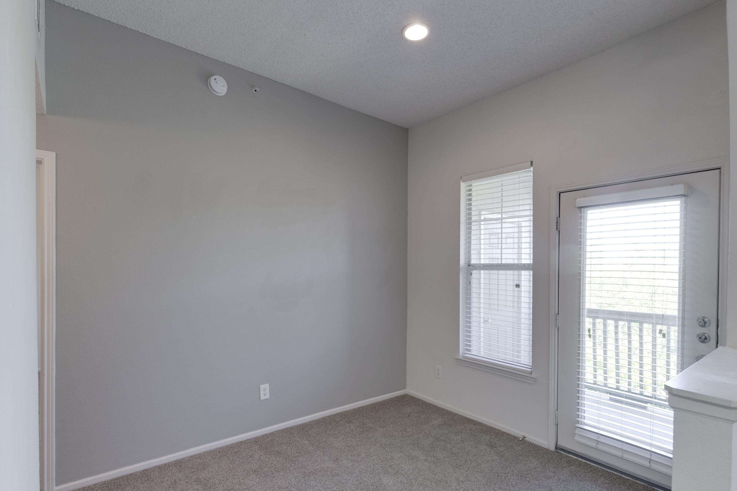 Empty room with gray walls and carpeted floor. A large window with white blinds lets in natural light, and a door with blinds leads to an outside balcony. The ceiling has a light fixture and a smoke detector. The space is bright and minimalistic, ready for furnishing.