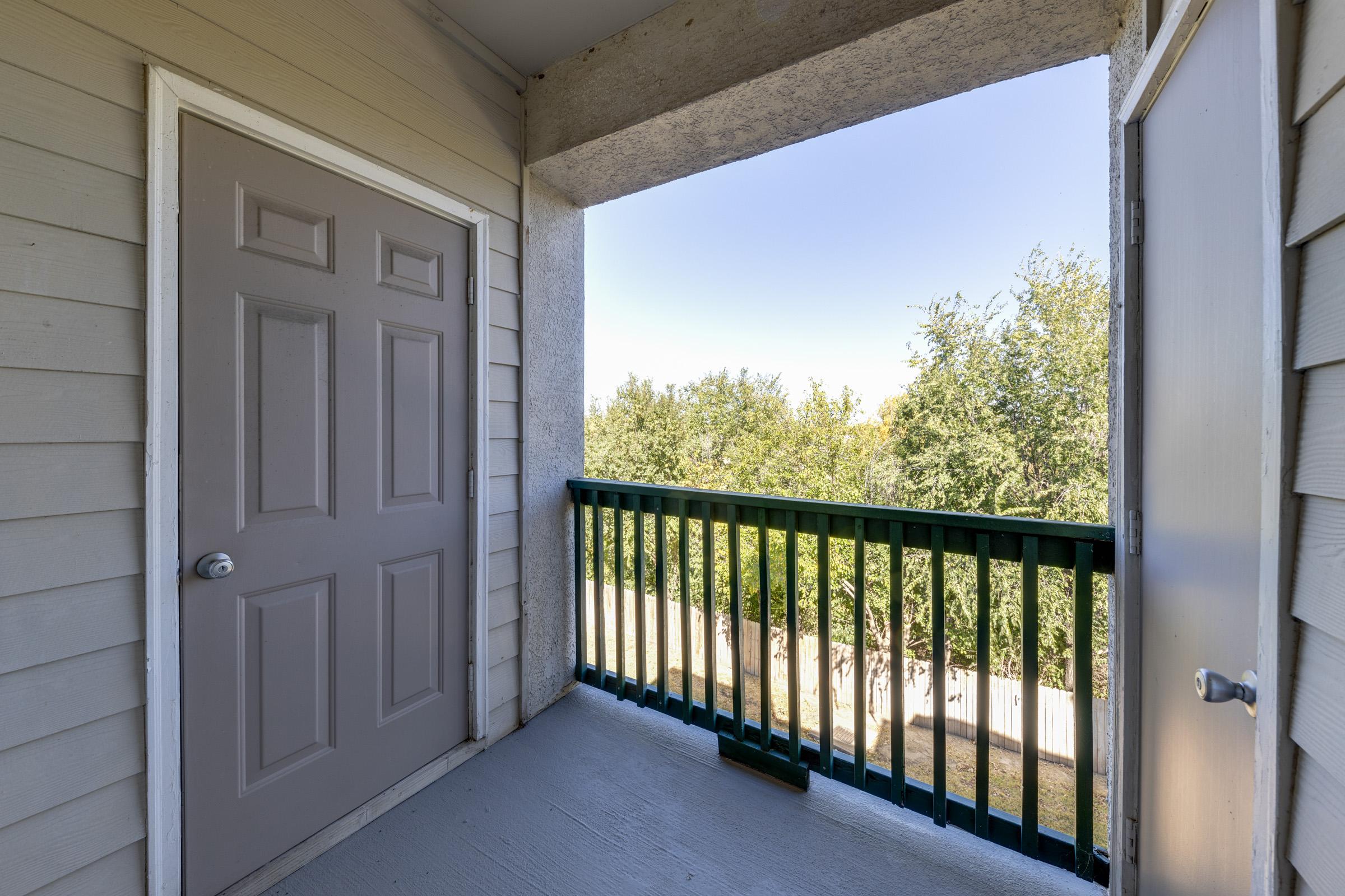 A small balcony with a light gray door leading indoors, surrounded by beige siding. The balcony has a green railing, with a view of trees outside in the distance and a clear blue sky overhead. A second door is visible on the right side leading to another space.