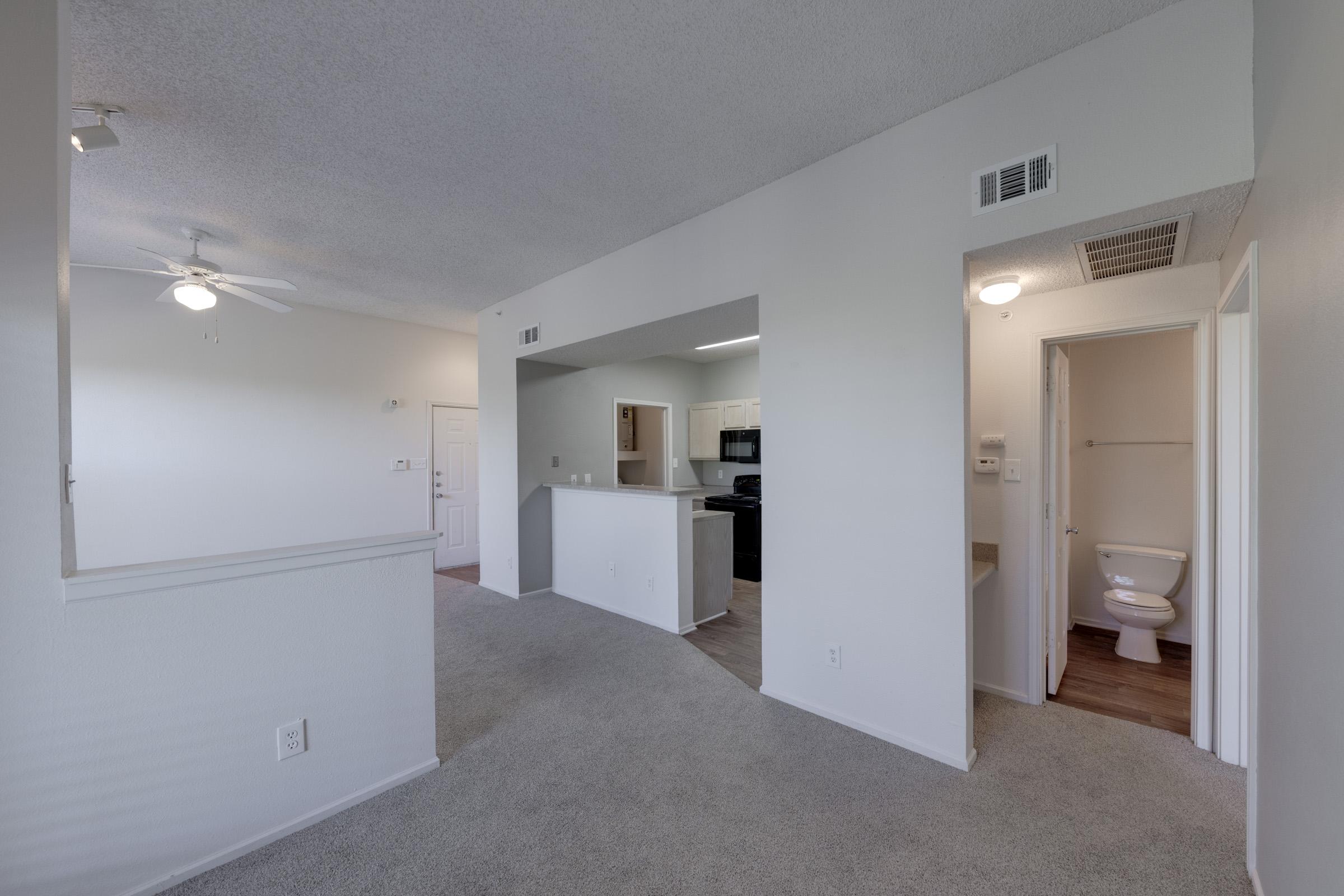 A spacious, well-lit interior of an apartment featuring a living area with carpet, a ceiling fan, and an open layout leading to a kitchen area with modern appliances. A door leads to a bathroom with a toilet, and there's a view of the entryway from the living space.