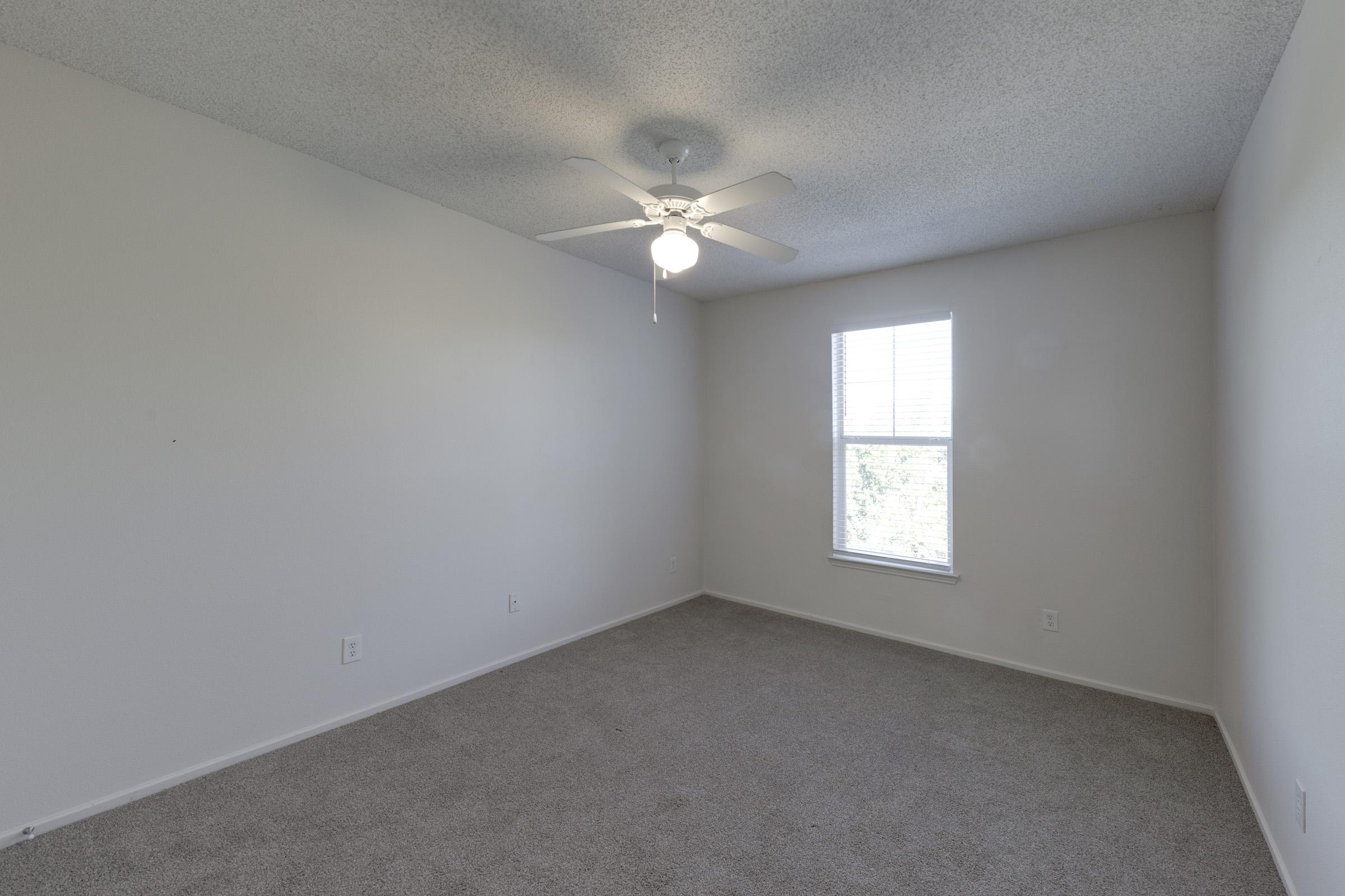 A spacious, empty room with light gray carpet, white walls, and a ceiling fan. There's one window allowing natural light to enter, casting a soft glow in the area. The room is unfurnished, creating a minimalist and open feel.