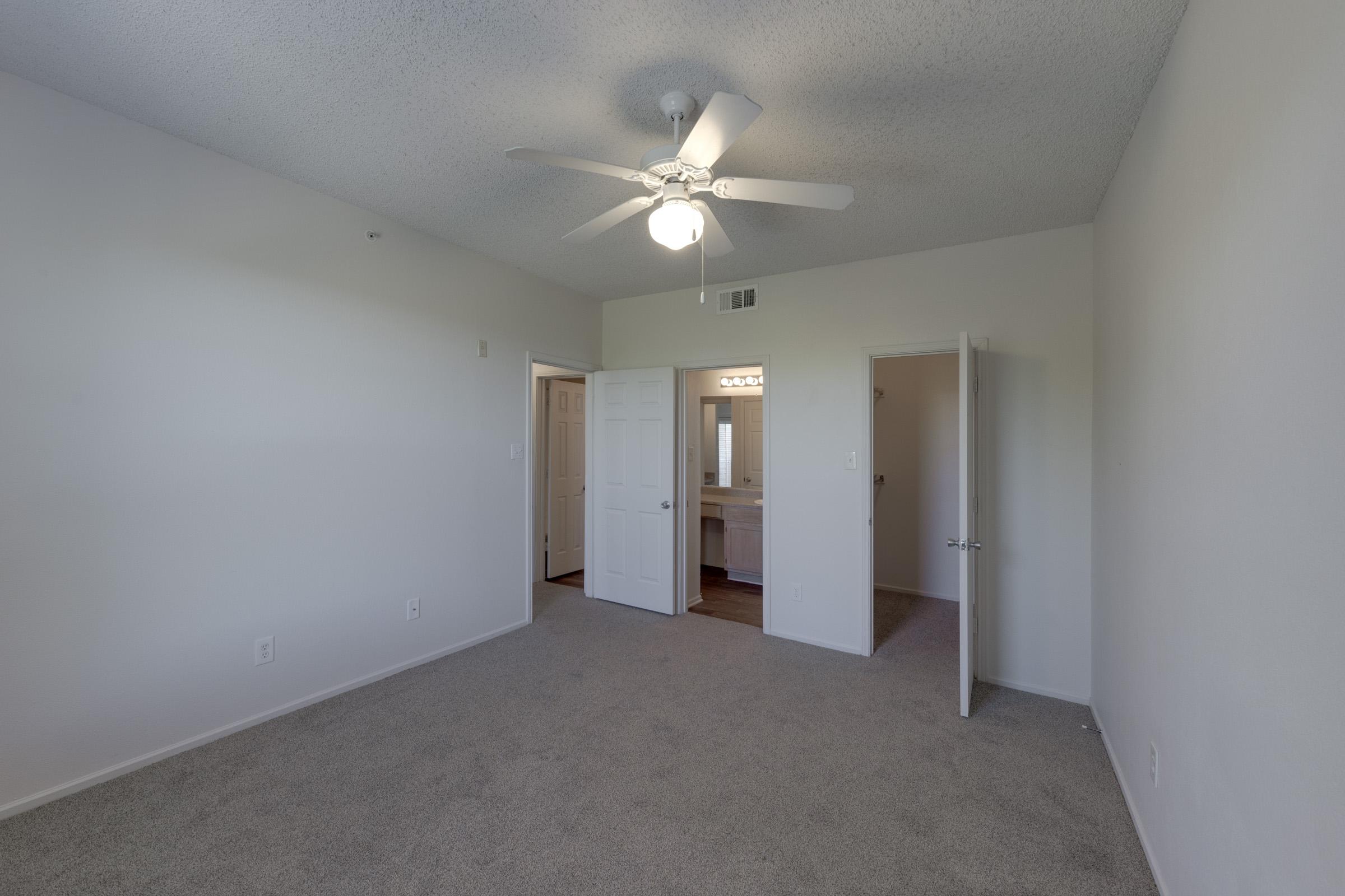 A small, empty room with light-colored walls and a textured ceiling. The floor is covered in beige carpet. A ceiling fan is installed in the center. Two doors are visible: one leads to a bathroom with lights visible through an open door, and the other is closed. The room is bright and airy.