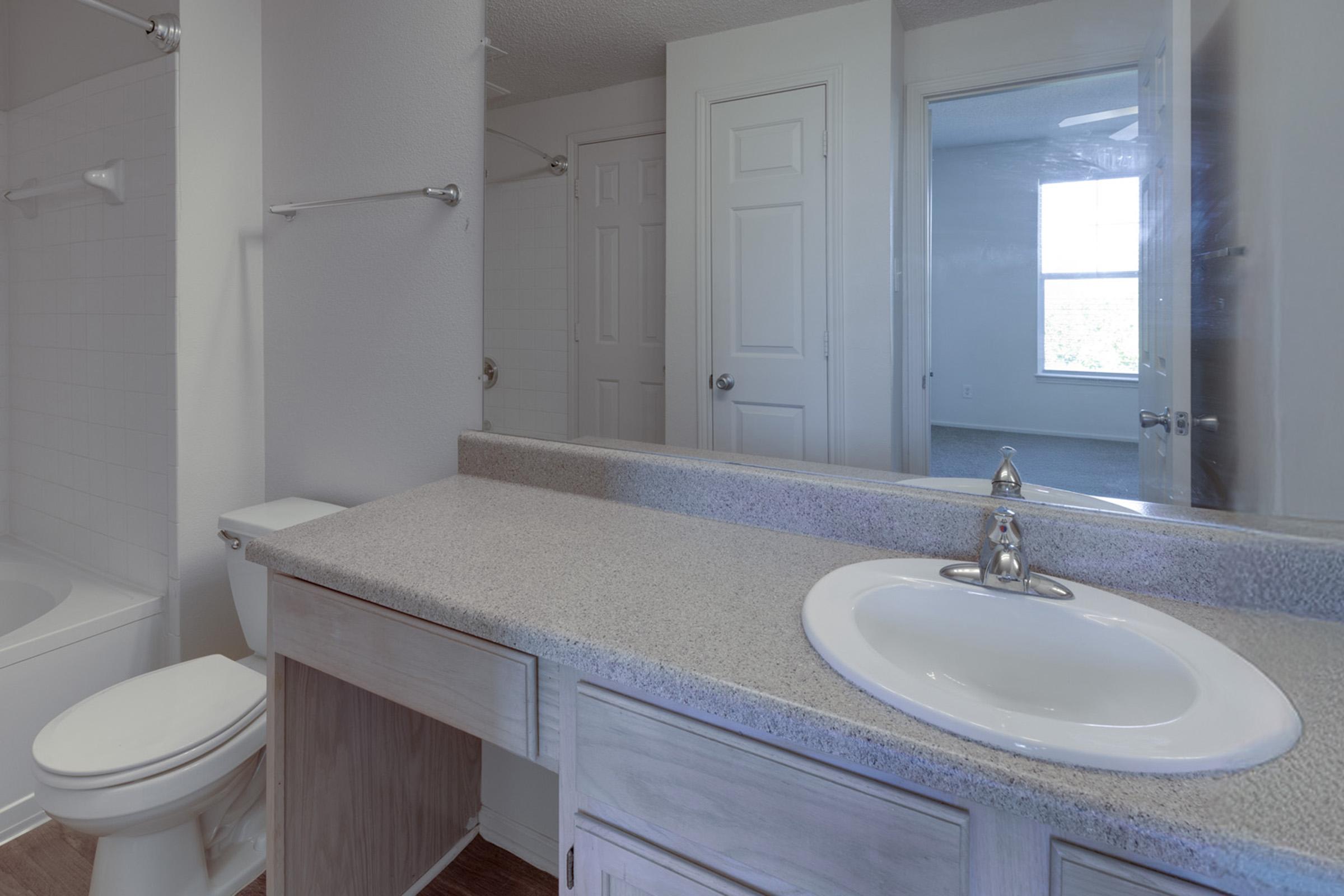 A modern bathroom featuring a white sink with a faucet on a light-colored countertop. There is a mirror above the sink, and a bathtub is visible on the left. A doorway leads to another room, and a window provides natural light, reflecting in the mirror and adding brightness to the space.