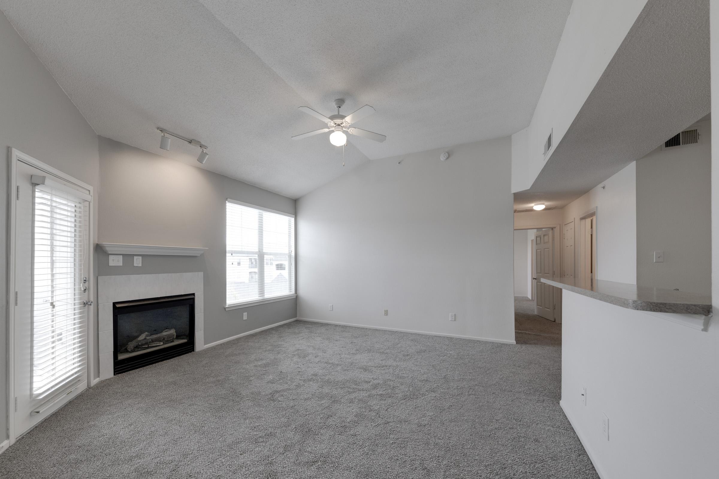 A spacious living room with light gray carpet, a ceiling fan, and a fireplace. There are large windows allowing natural light, along with a door leading outside. The room has white walls and a vaulted ceiling, creating an open and airy feel. An adjoining kitchen area is visible in the background.