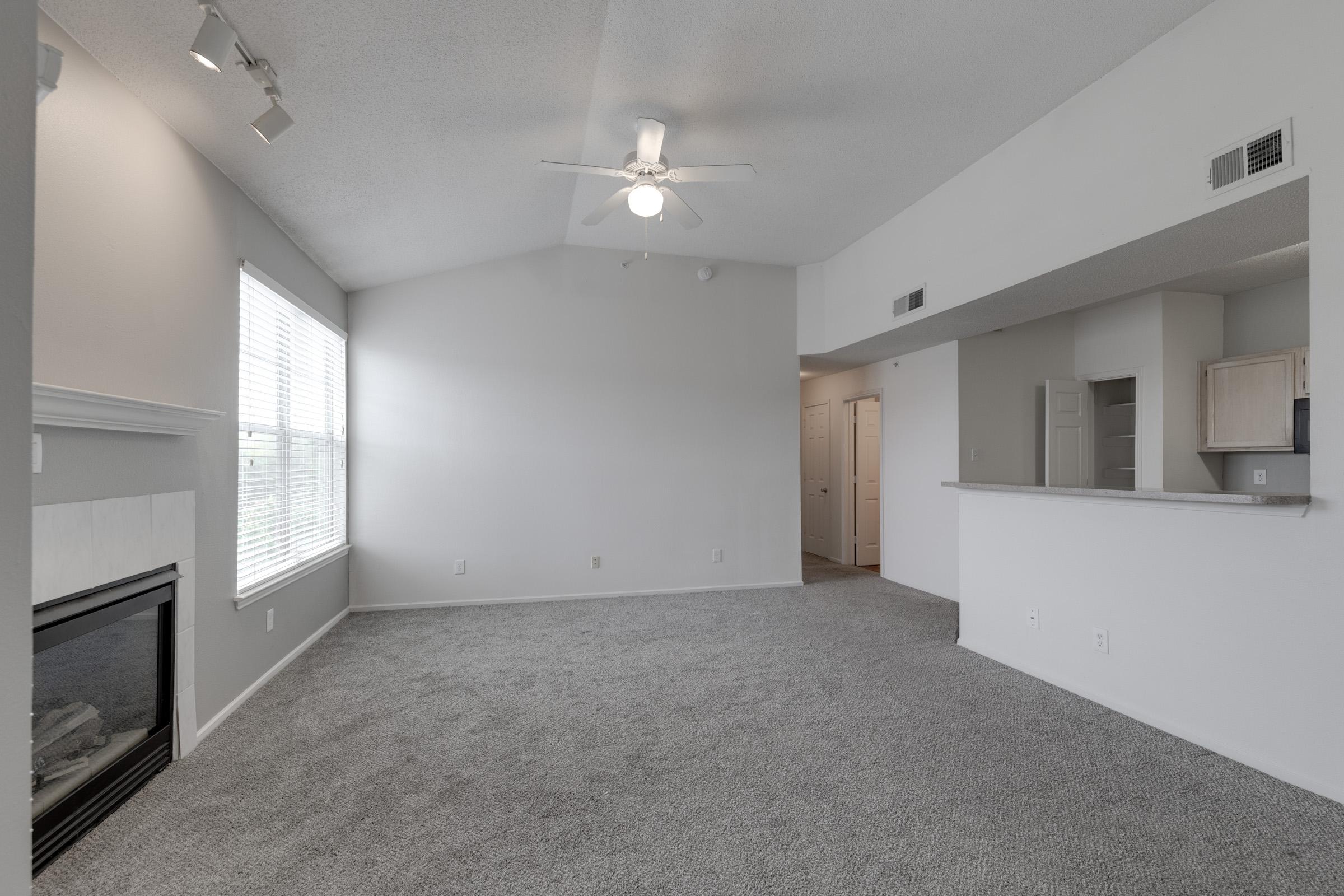 A spacious, carpeted living room with a ceiling fan, a modern fireplace, and large windows allowing natural light. The room features neutral-colored walls and a partially open kitchen area with light cabinetry, creating a welcoming atmosphere.