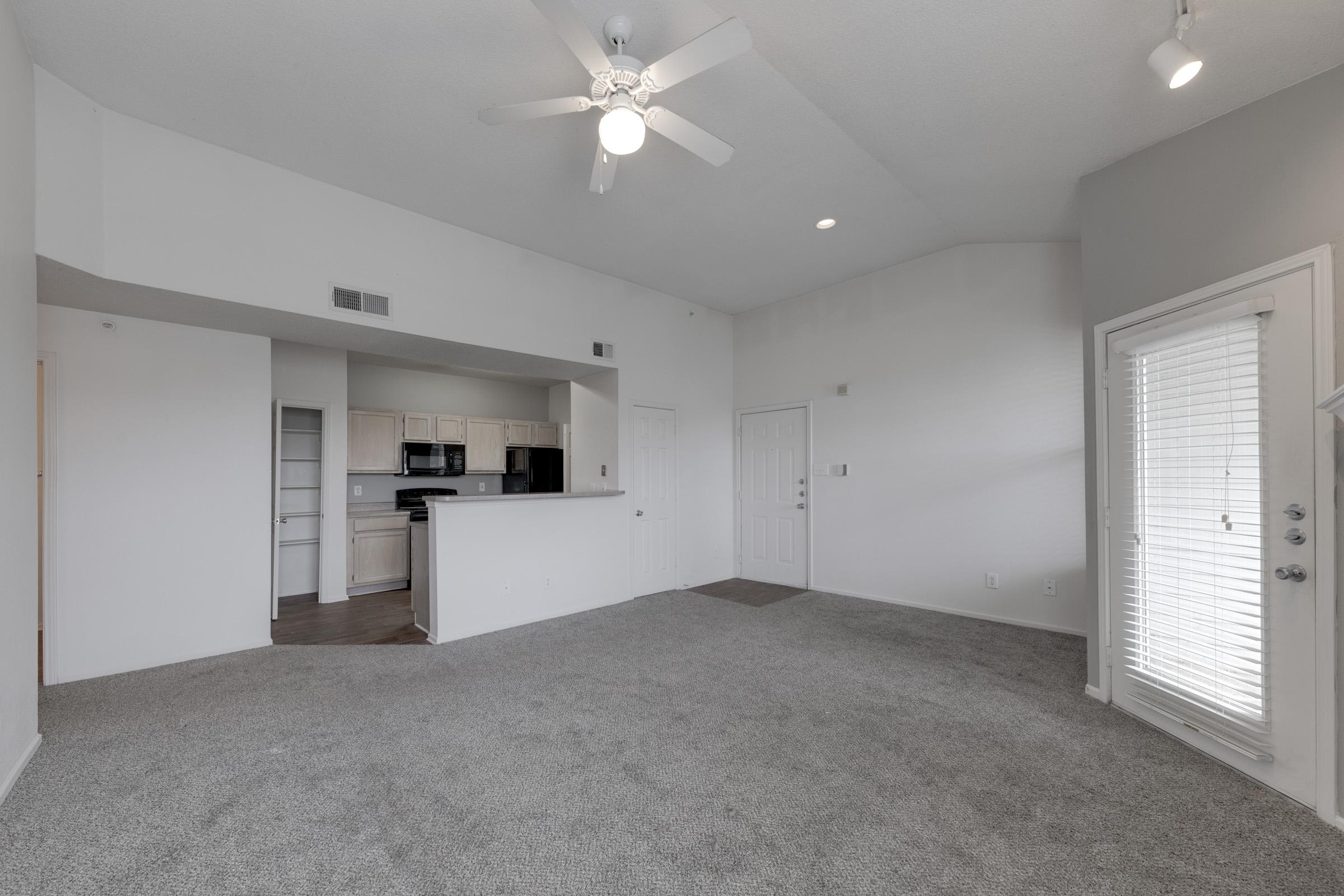 Spacious living area with light gray carpet, ceiling fan, and tall ceilings. An open kitchen area features light cabinetry and countertops. A door leads to a small patio with windows, allowing natural light into the space. Modern design with neutral colors for a cozy atmosphere.